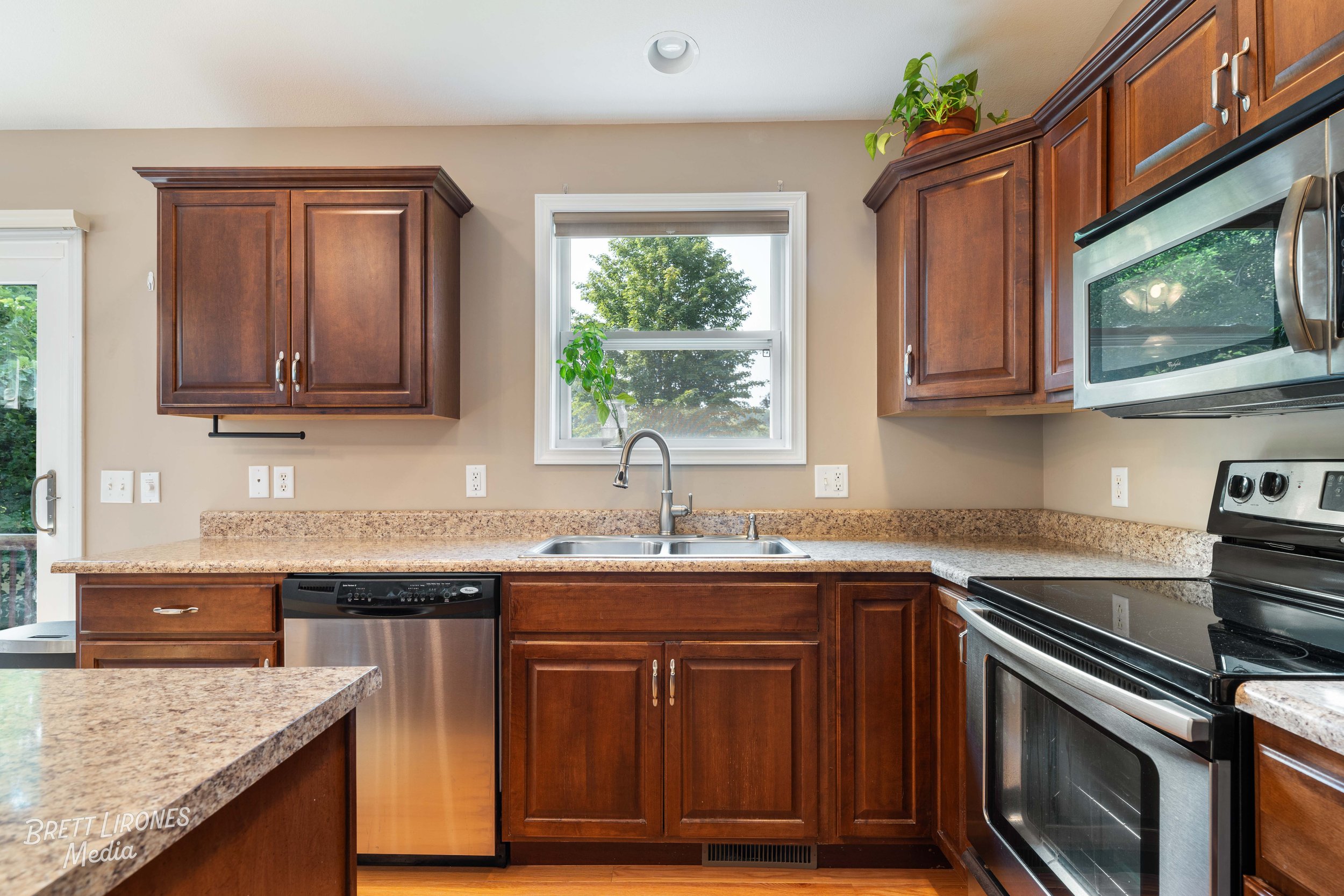 Kitchen with brown cabinets, granite countertops, stainless steel appliances, a window above the sink, and potted plants on top of the cabinets.
