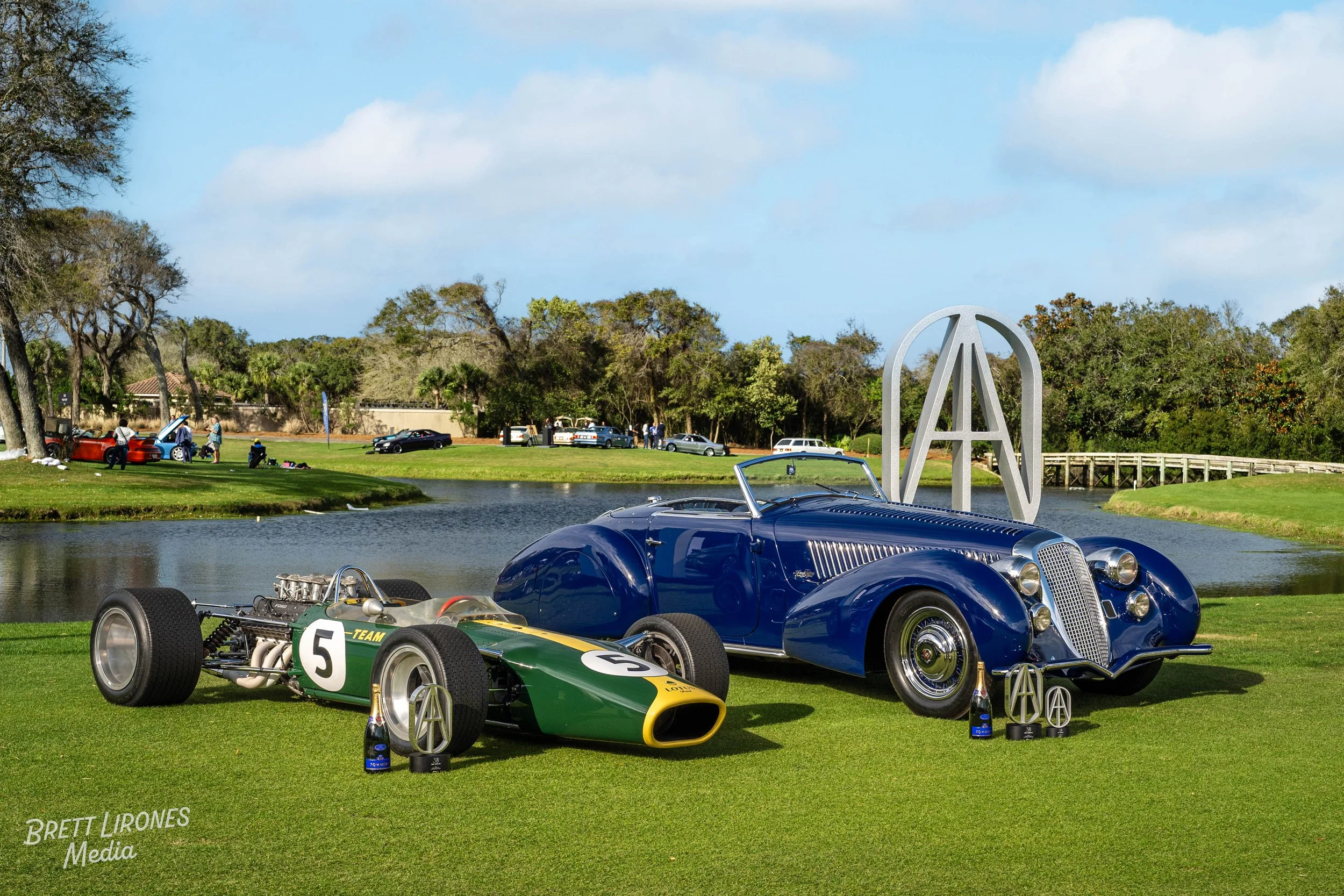 Vintage blue convertible car and a green and yellow race car on grass near a body of water, with an 'Aston Martin' logo sculpture and trophies in front, and people and cars in the background.