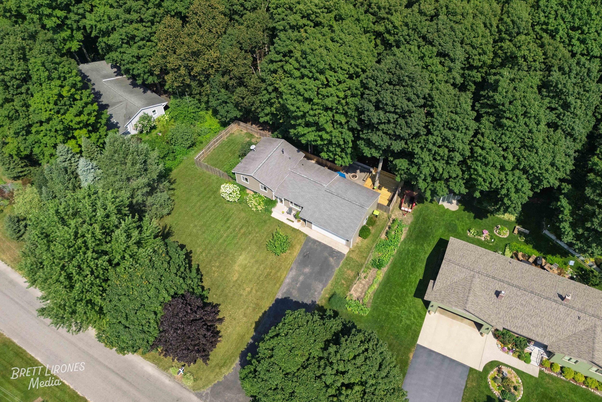 Aerial view of a suburban neighborhood featuring a house with a gray roof, a large backyard with a deck and patio, green lawns, and surrounded by tall trees.