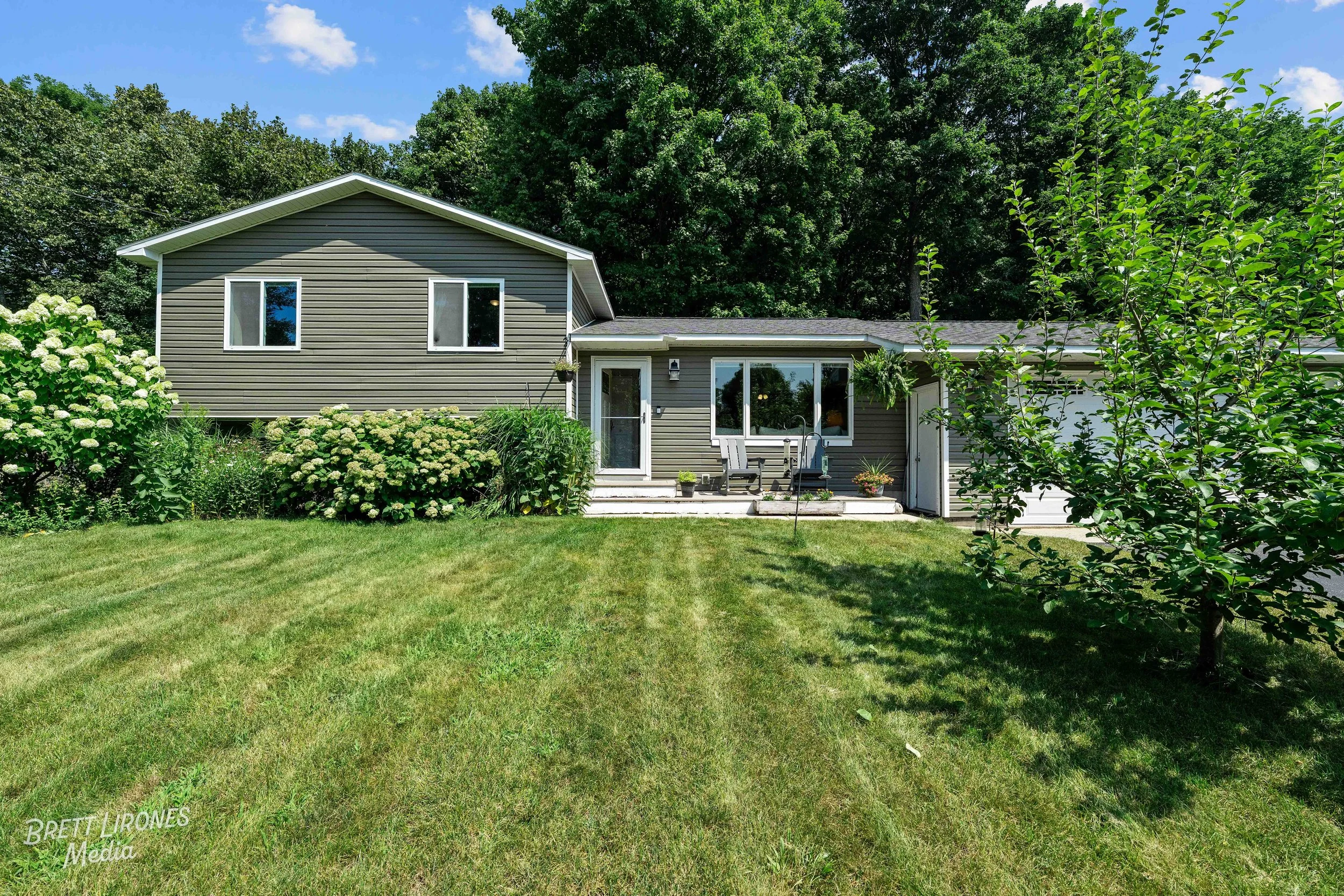 A backyard with a grassy lawn, a small patio with outdoor furniture, a greenhouse, and a gray house with sliding glass doors. There are trees and shrubs surrounding the yard, and a small tree on the right side.