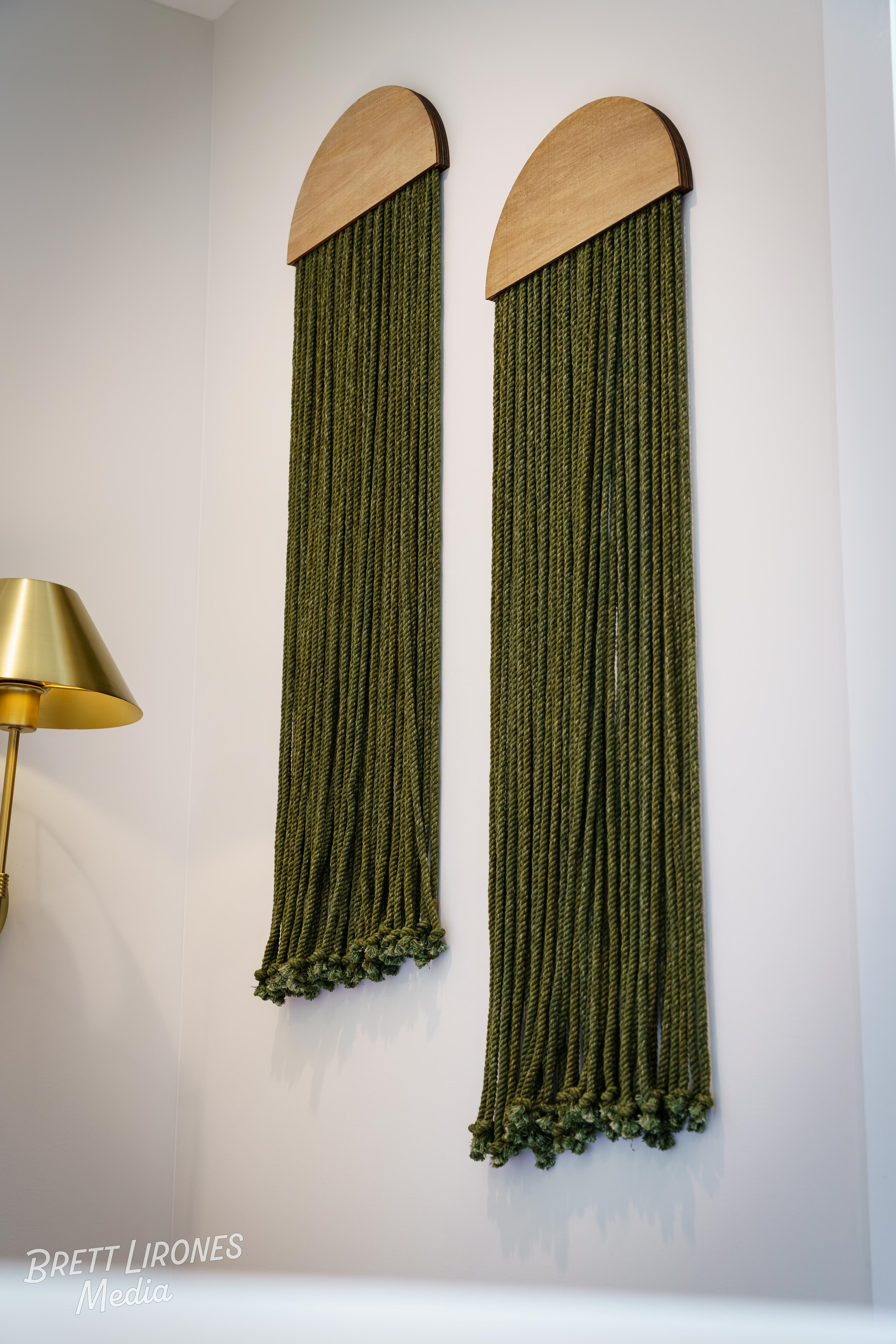 Two green velvet curtains with wooden half-circle tops and knotted tassels at the bottom, hanging on a white wall, with a gold-colored lamp visible at the left edge.