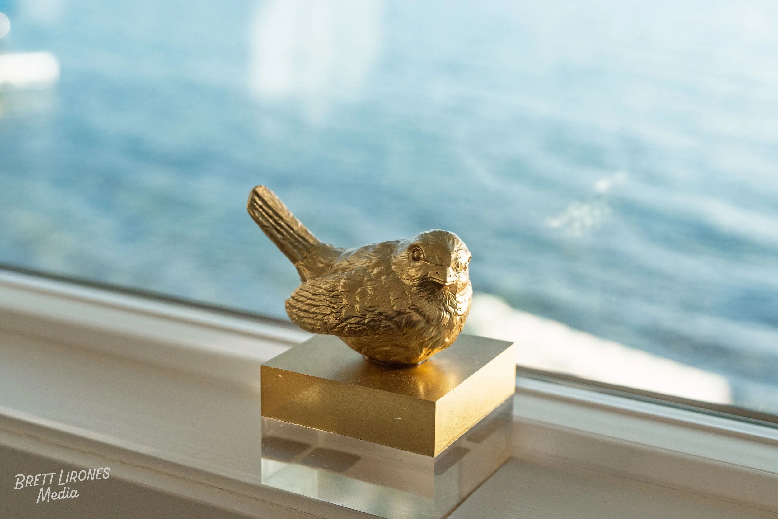 A decorative sculpture of a bird with a metallic finish, positioned on a square base, placed on a windowsill with a body of water visible outside the window.