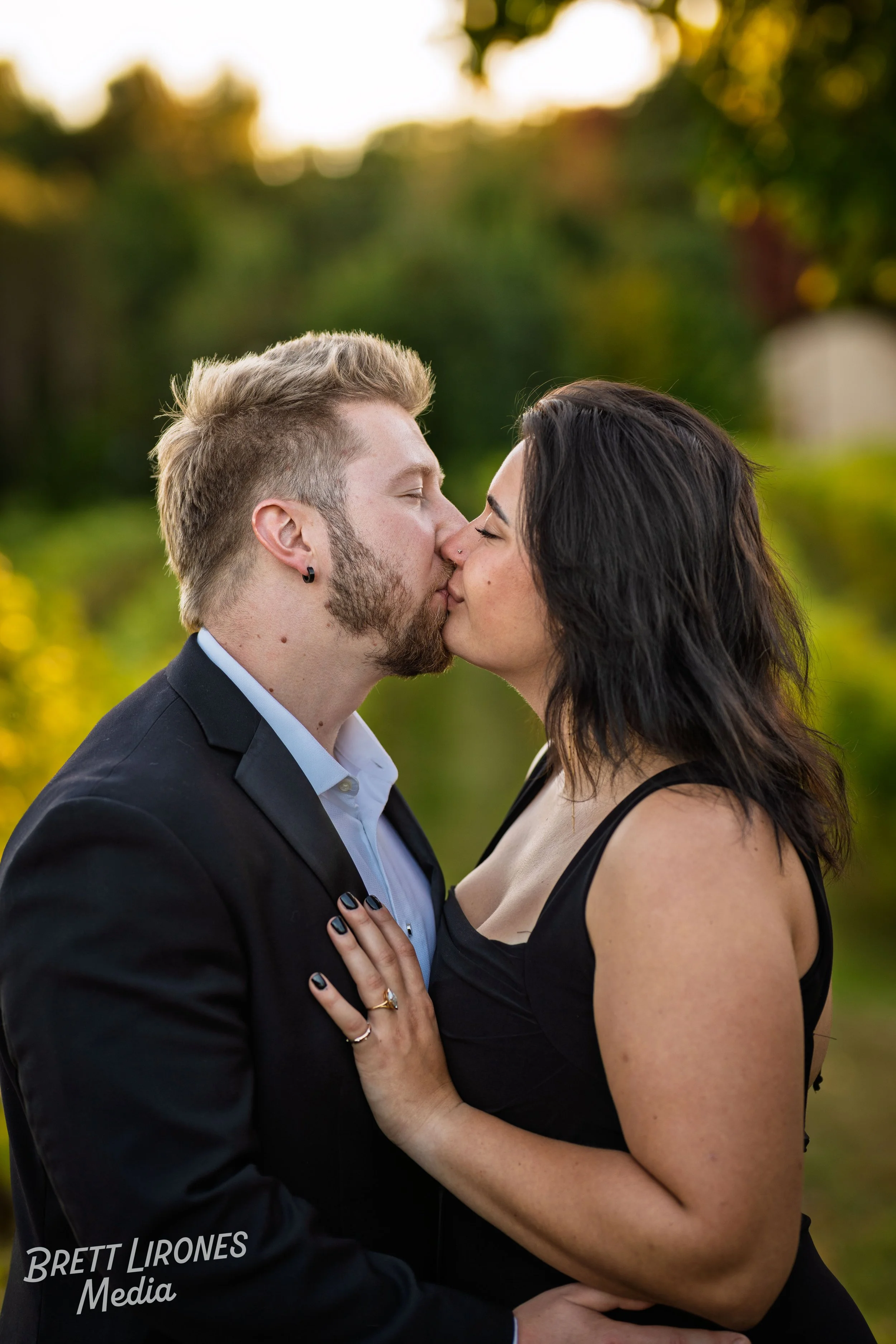 A couple kissing outdoors during sunset with trees in the background.