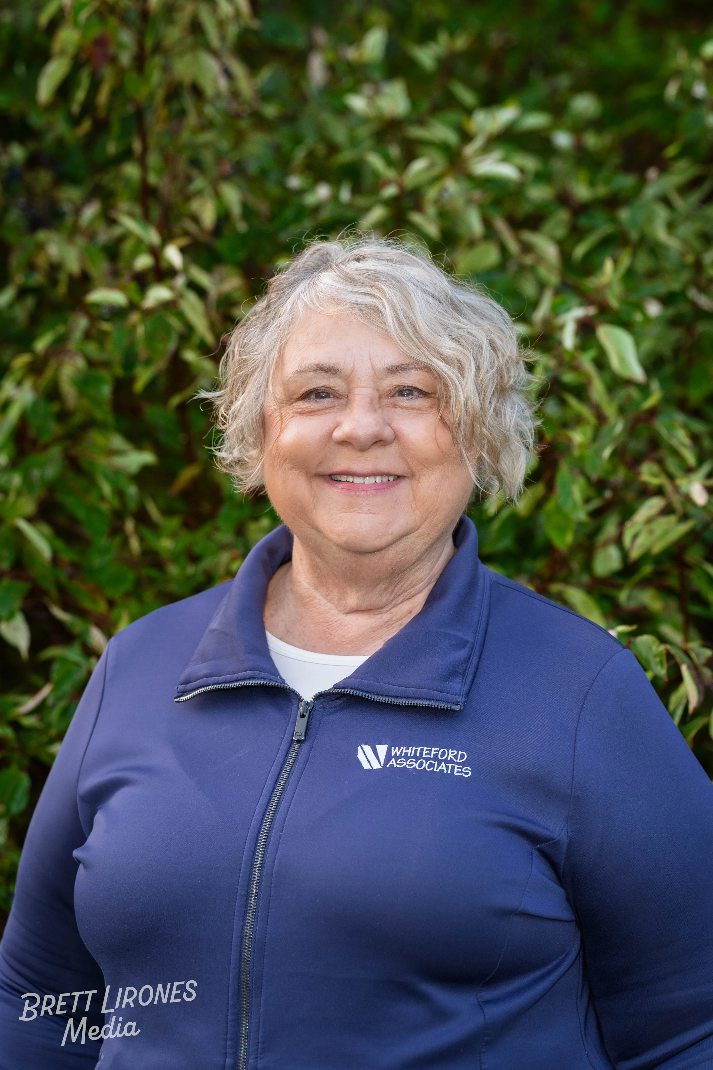 A smiling woman with short, curly, gray hair standing outdoors in front of green foliage, wearing a navy blue jacket with 'Whiteford Associates' logo.