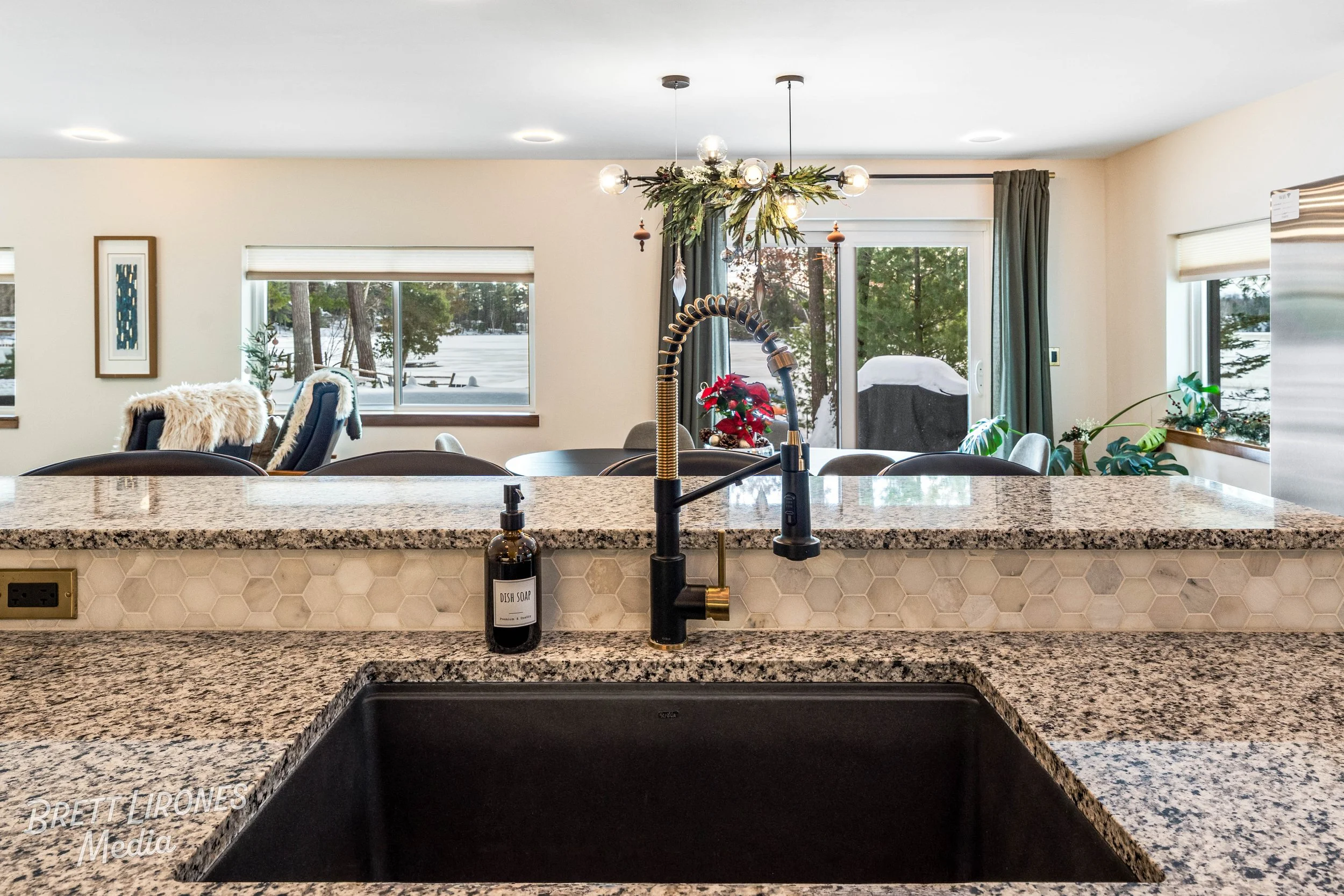 Kitchen with granite countertops, a black sink, a gold faucet, and a soap dispenser. In the background, there is a dining area with a round table, chairs, and large windows showing a snowy outdoor landscape with trees.