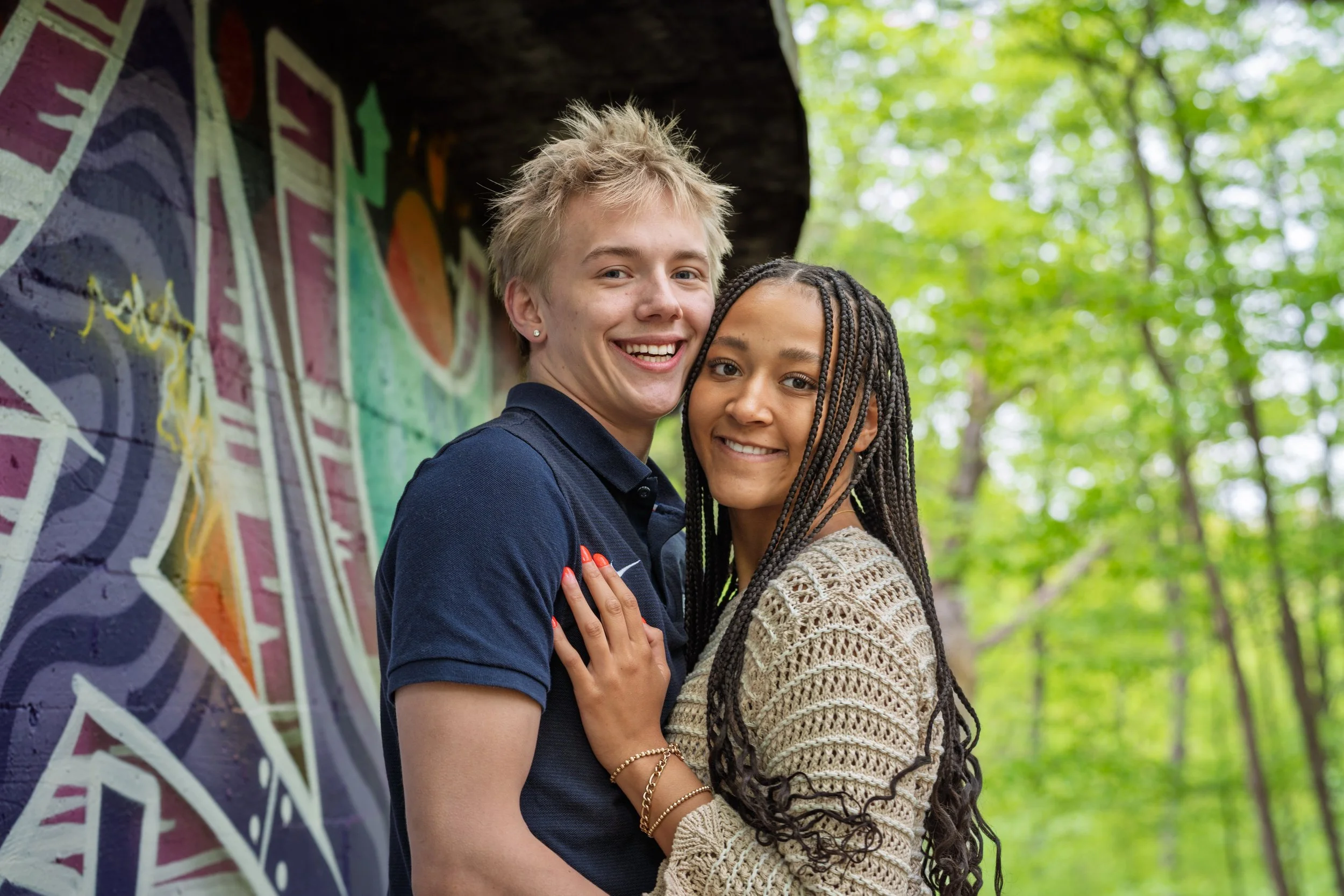 A smiling couple hugging outdoors near colorful graffiti, with green trees in the background.