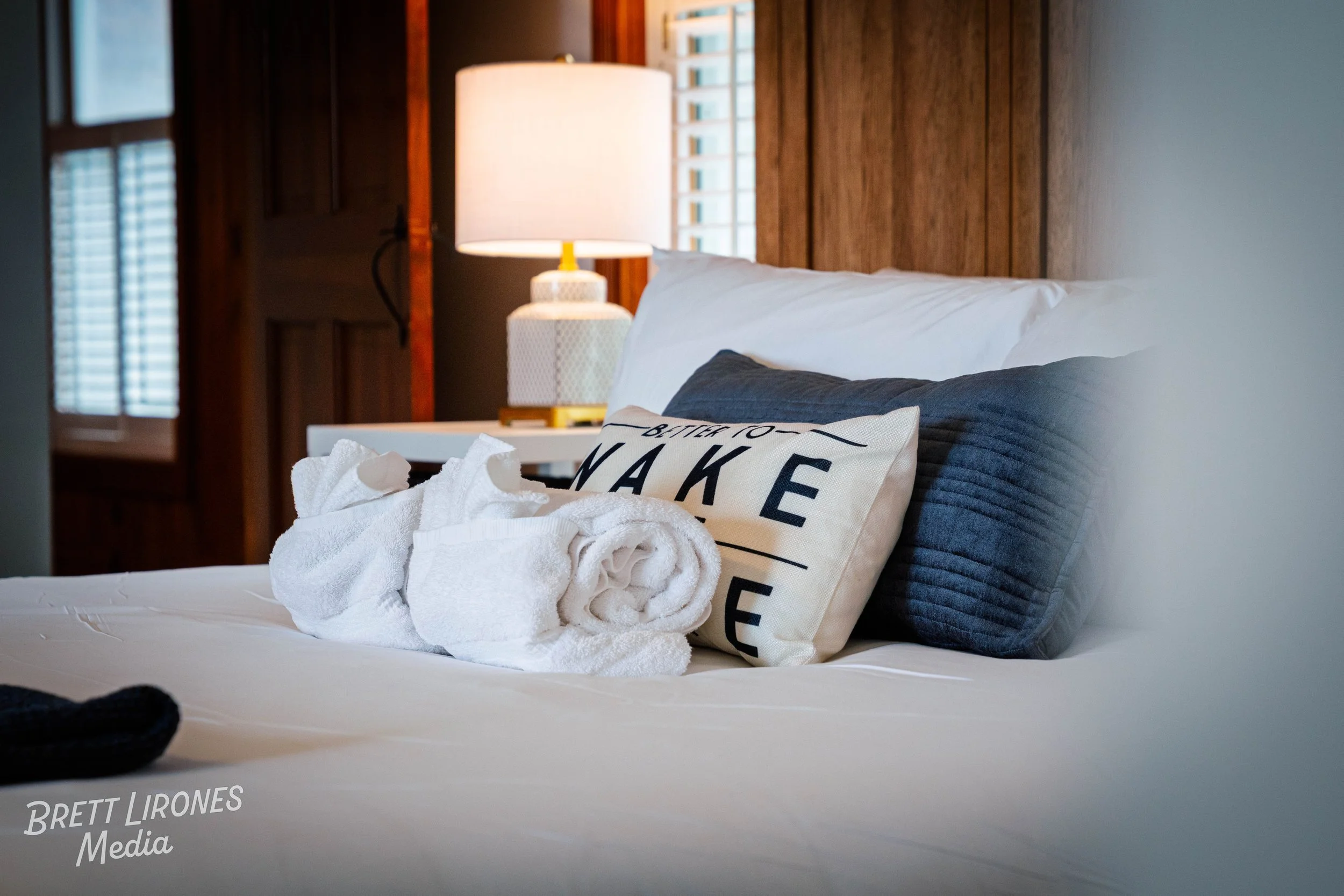 Close-up of a neatly made bed with a blue pillow, white pillow with black text, and towel sculptures on top, in a cozy bedroom with wooden walls and a table lamp.