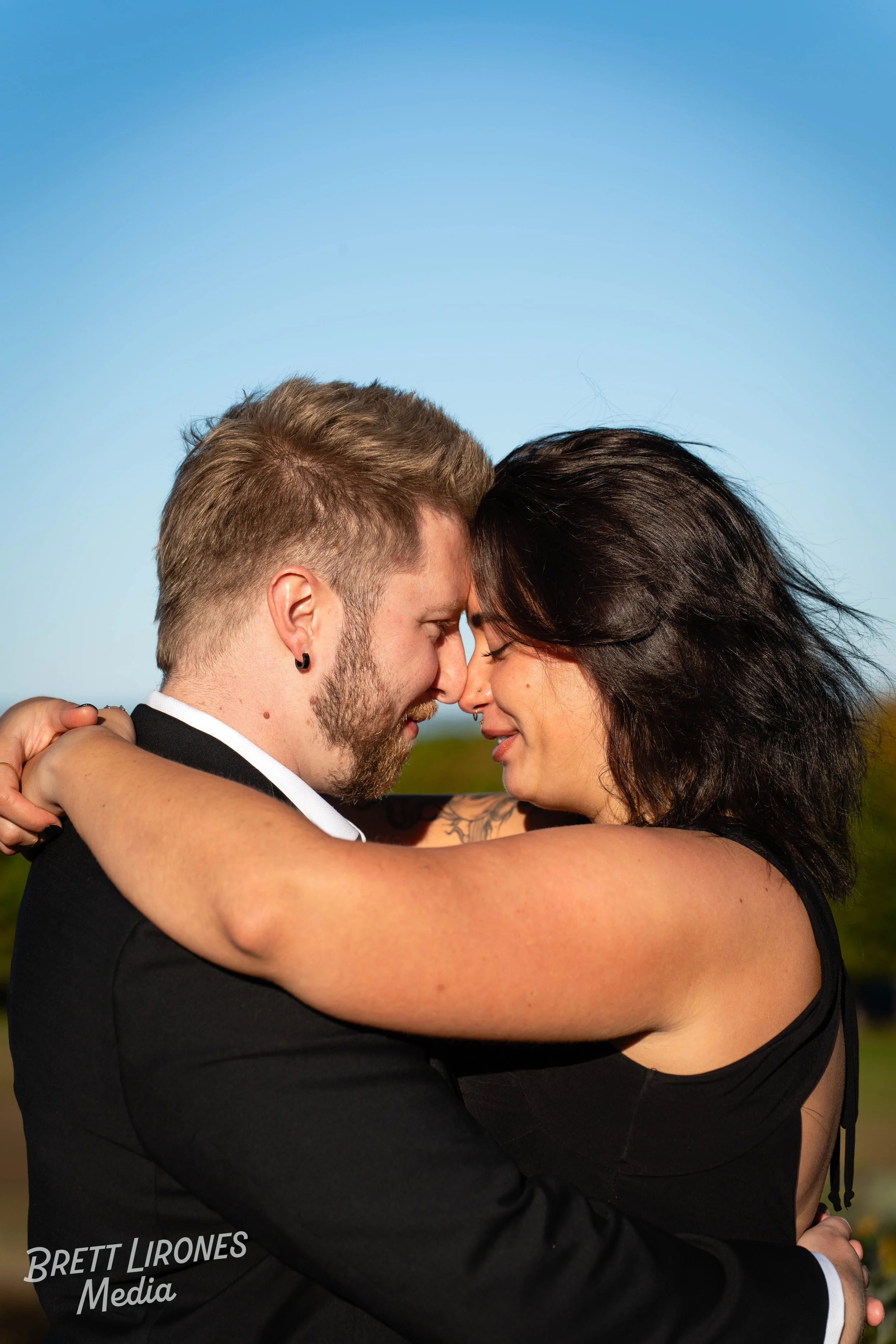 A couple embracing outdoors with foreheads and noses touching, eyes closed, in a loving moment. The man has short hair and a beard, and the woman has shoulder-length dark hair and tattoos on her arms. They are dressed nicely, with the man in a black 