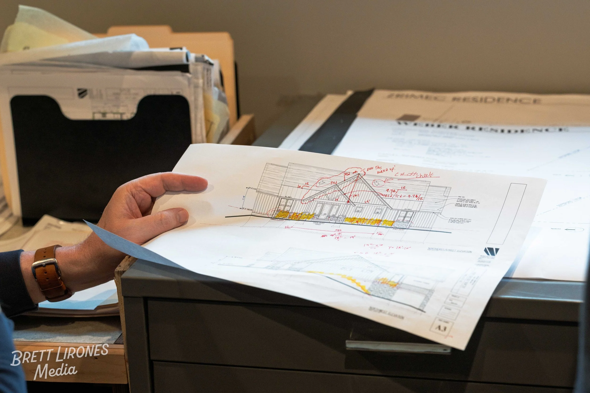 Person holding architectural blueprint of a house with notes, on a desk with files and plans, in an office setting.