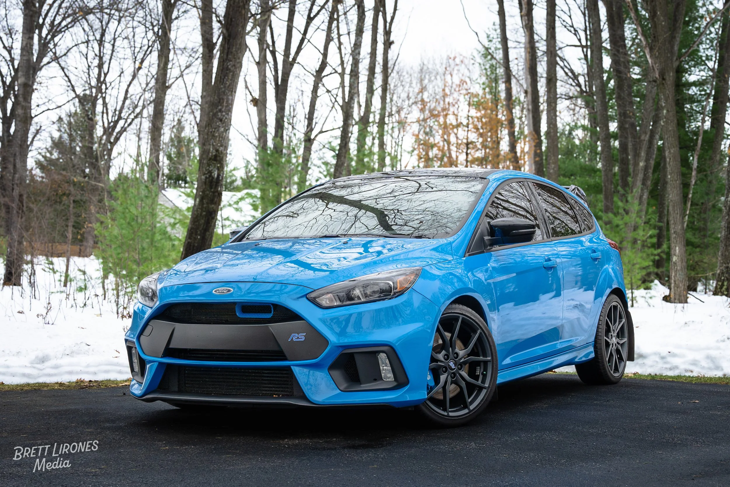 A blue Ford Focus RS parked on a dark asphalt driveway with snow and leafless trees in the background.