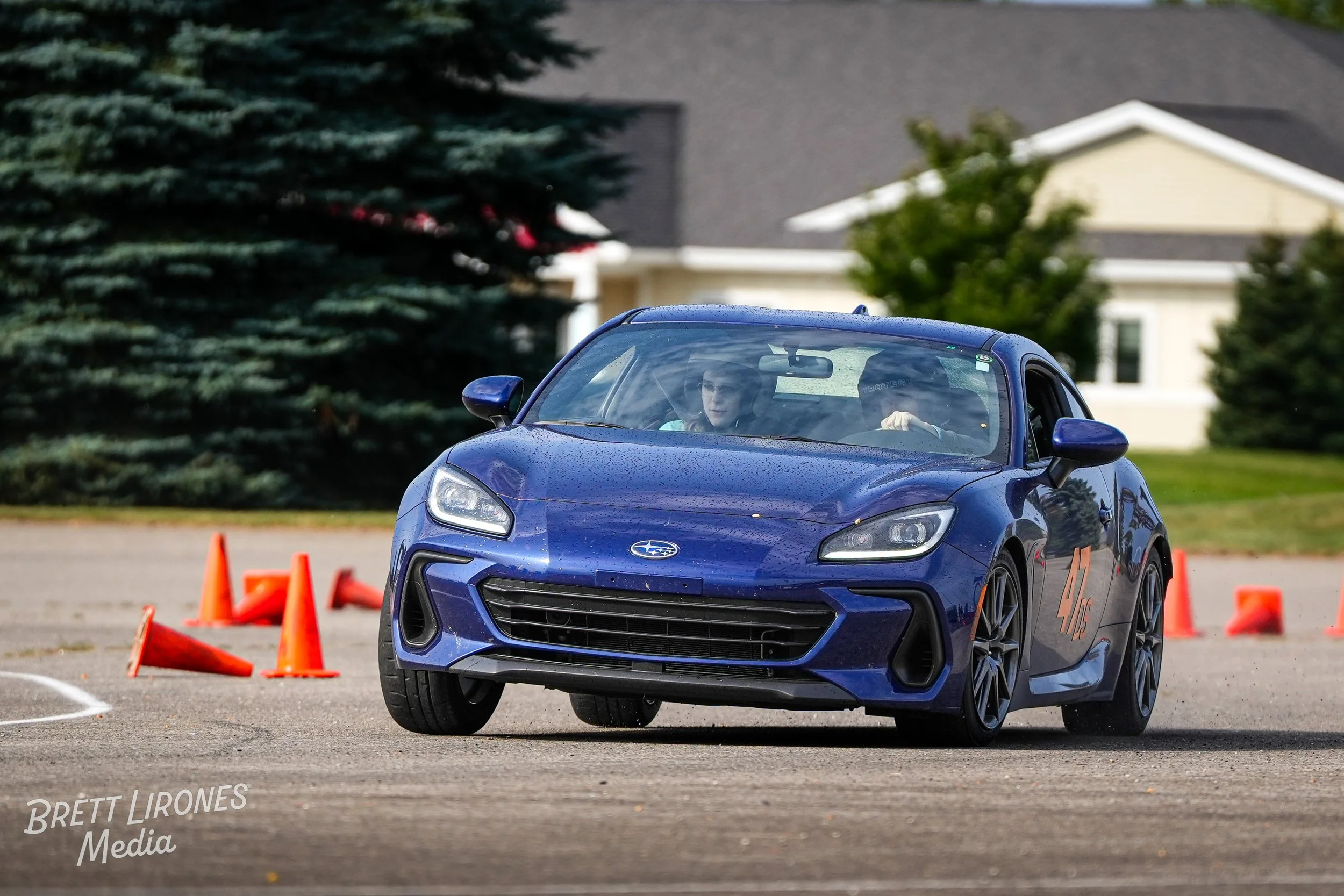 A blue Subaru BRZ sports car navigating through orange traffic cones on a paved course with two drivers inside during a driving event. The car has the number 44 printed on the side.