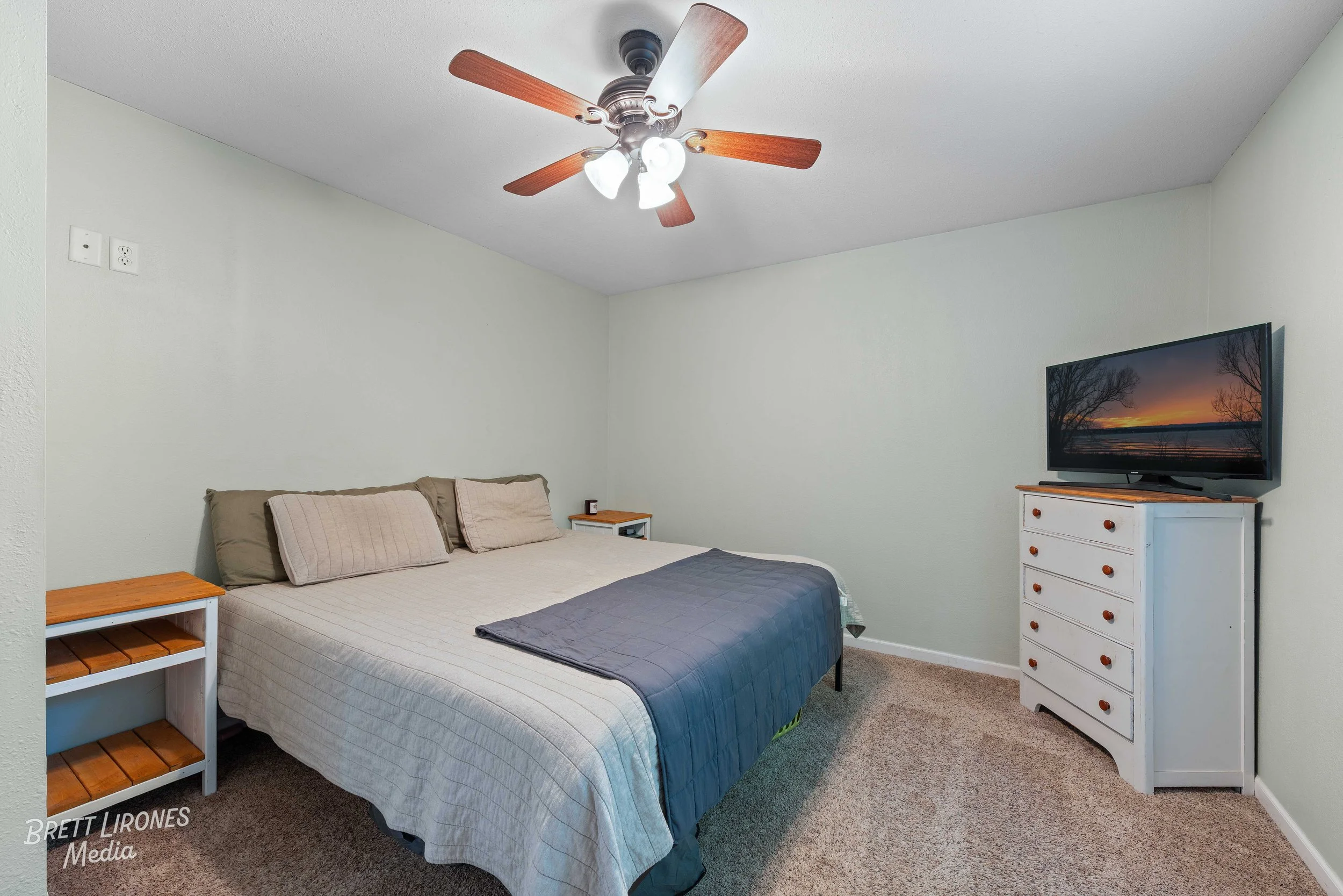 A bedroom with a bed, two nightstands, and a white dresser with a TV on top, beige carpet, a ceiling fan, and light green walls.