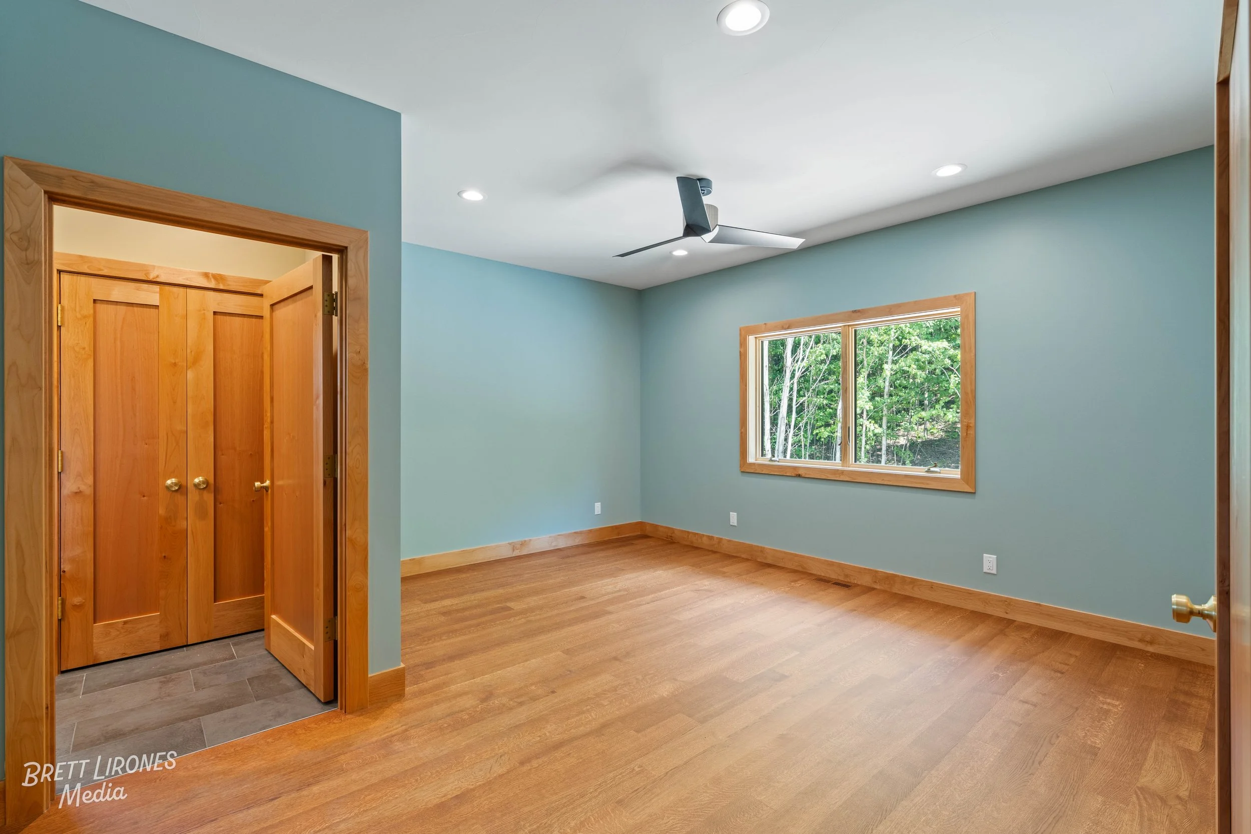 Empty bedroom with hardwood floors, light blue walls, a window showing trees outside, a ceiling fan, and a closet with wooden double doors.