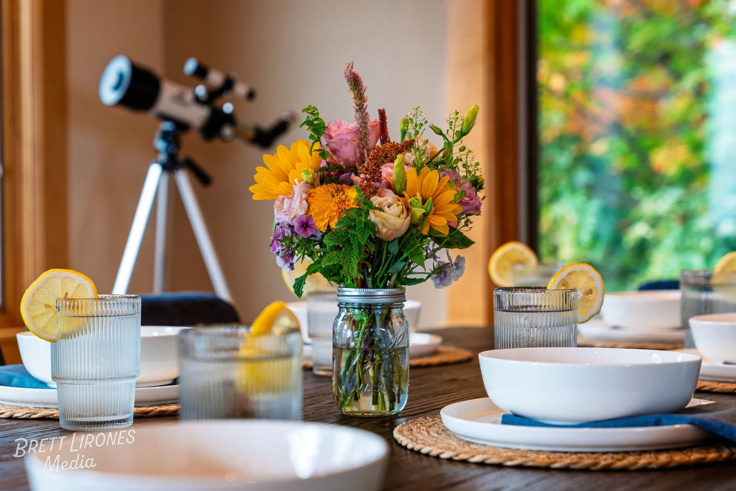 A dining table set with white bowls, glasses with lemon slices, and a centerpiece of colorful flowers in a glass jar, with a telescopic telescope in the background and a window showing green trees outside.