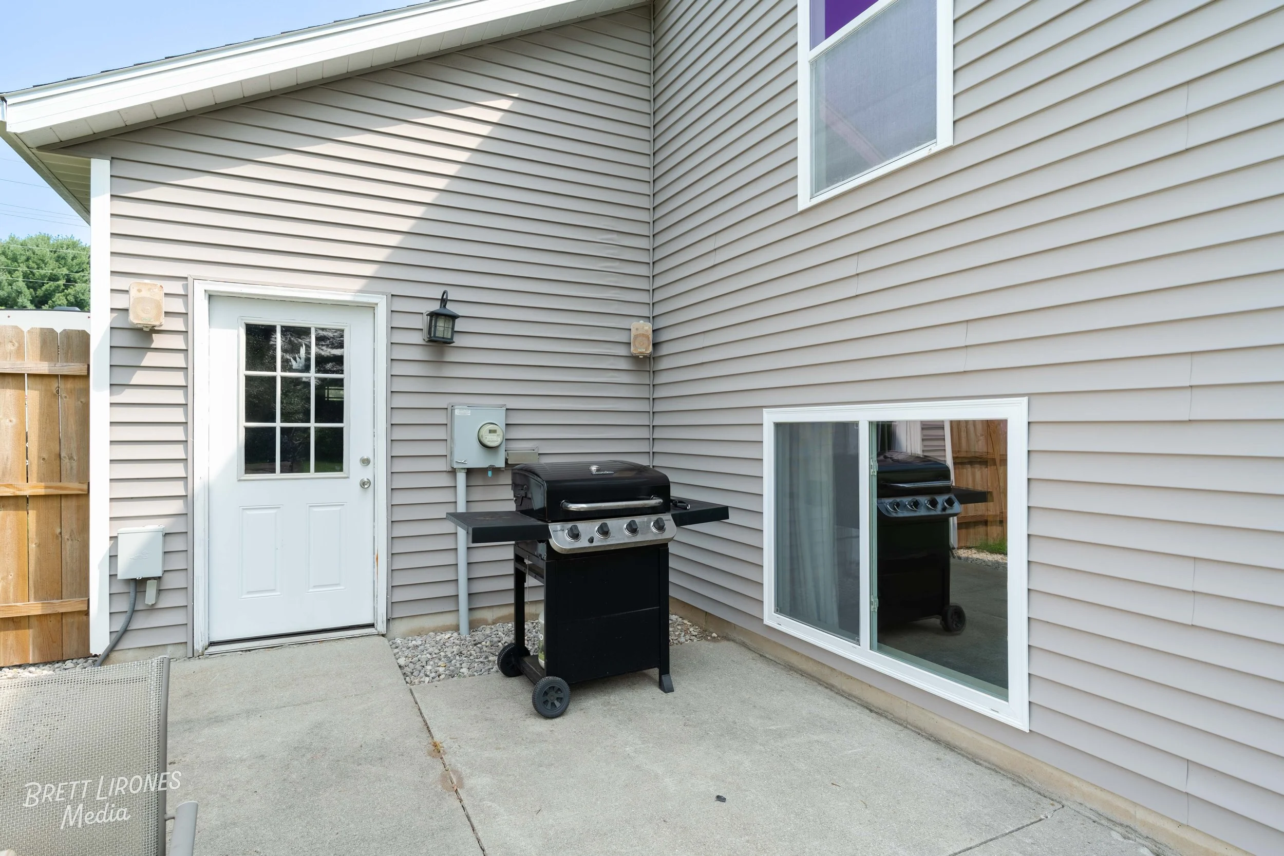 Back patio of a house with beige vinyl siding, a white door with glass panes, a sliding glass window, a gas grill, and a wooden privacy fence.
