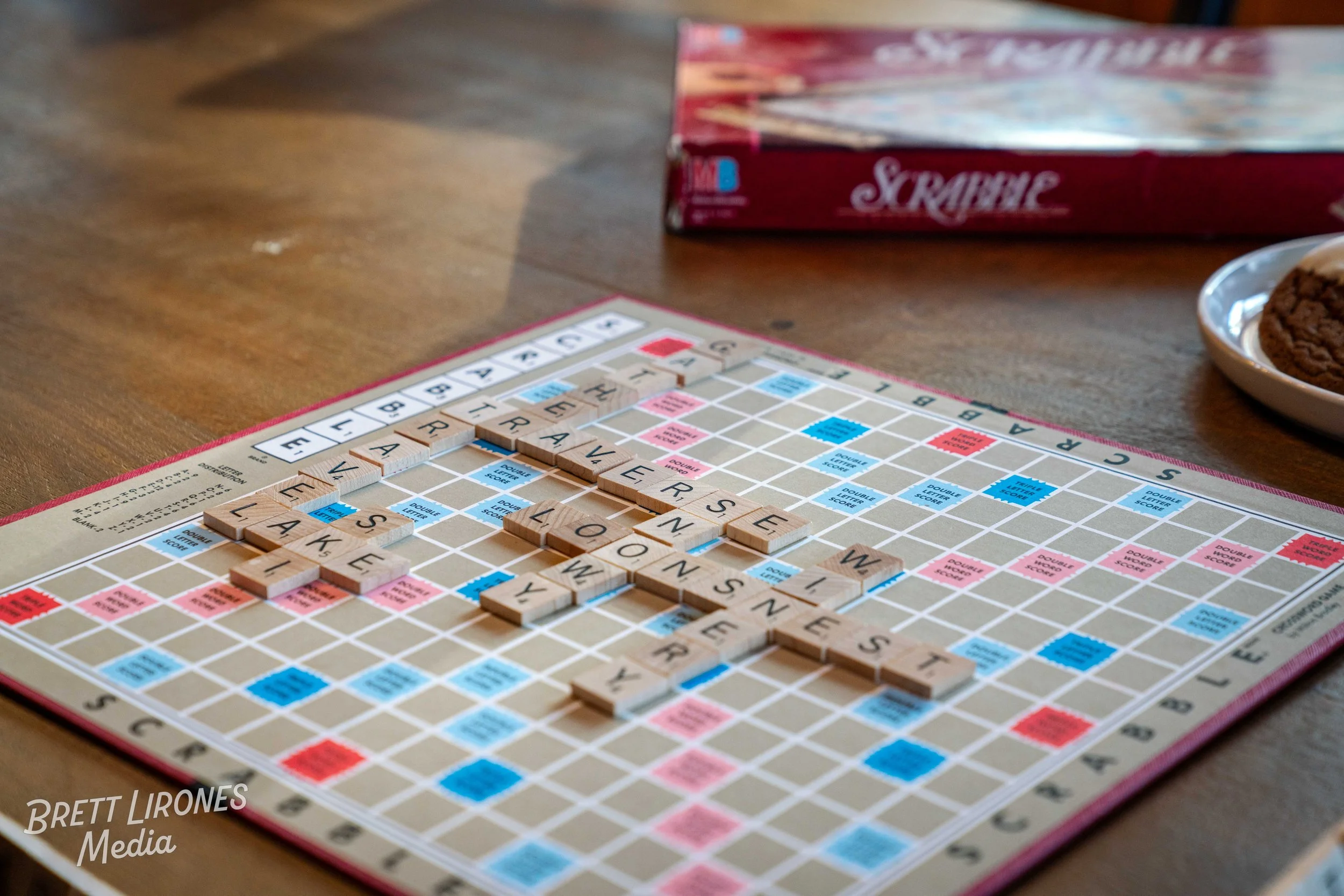 A Scrabble board game with words like 'FEEL' and 'LOVE' played on a wooden table, with a box of Scrabble and a plate with cookies nearby.
