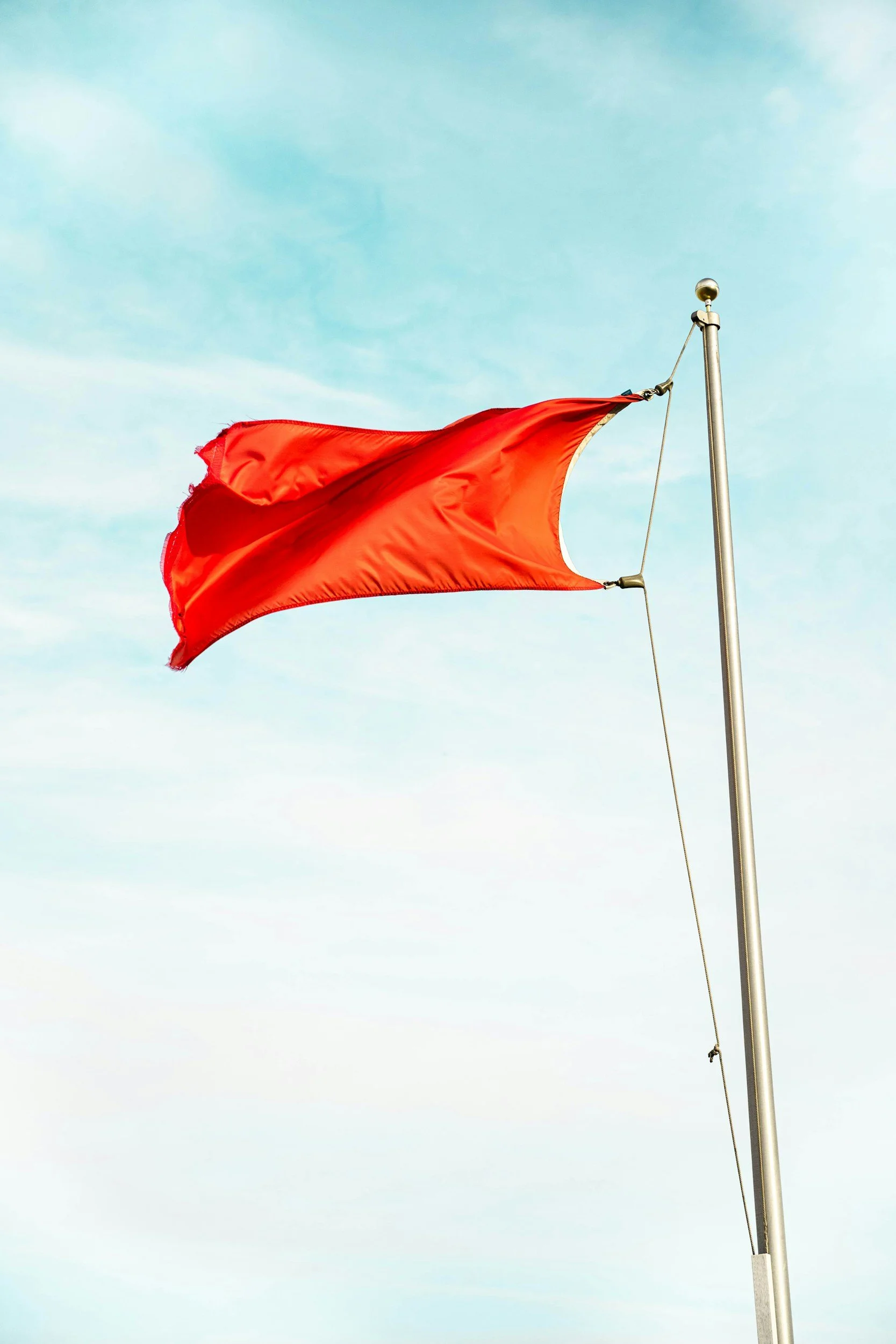 A red flag flying on a flagpole against a partly cloudy sky.