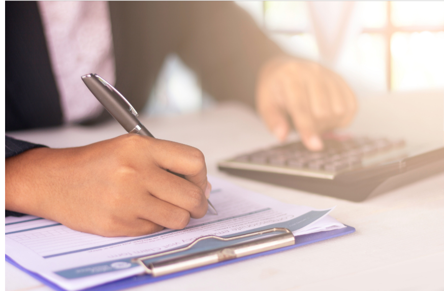 Close-up of a person's hand writing on a document with a pen, while another hand types on a calculator in the background.