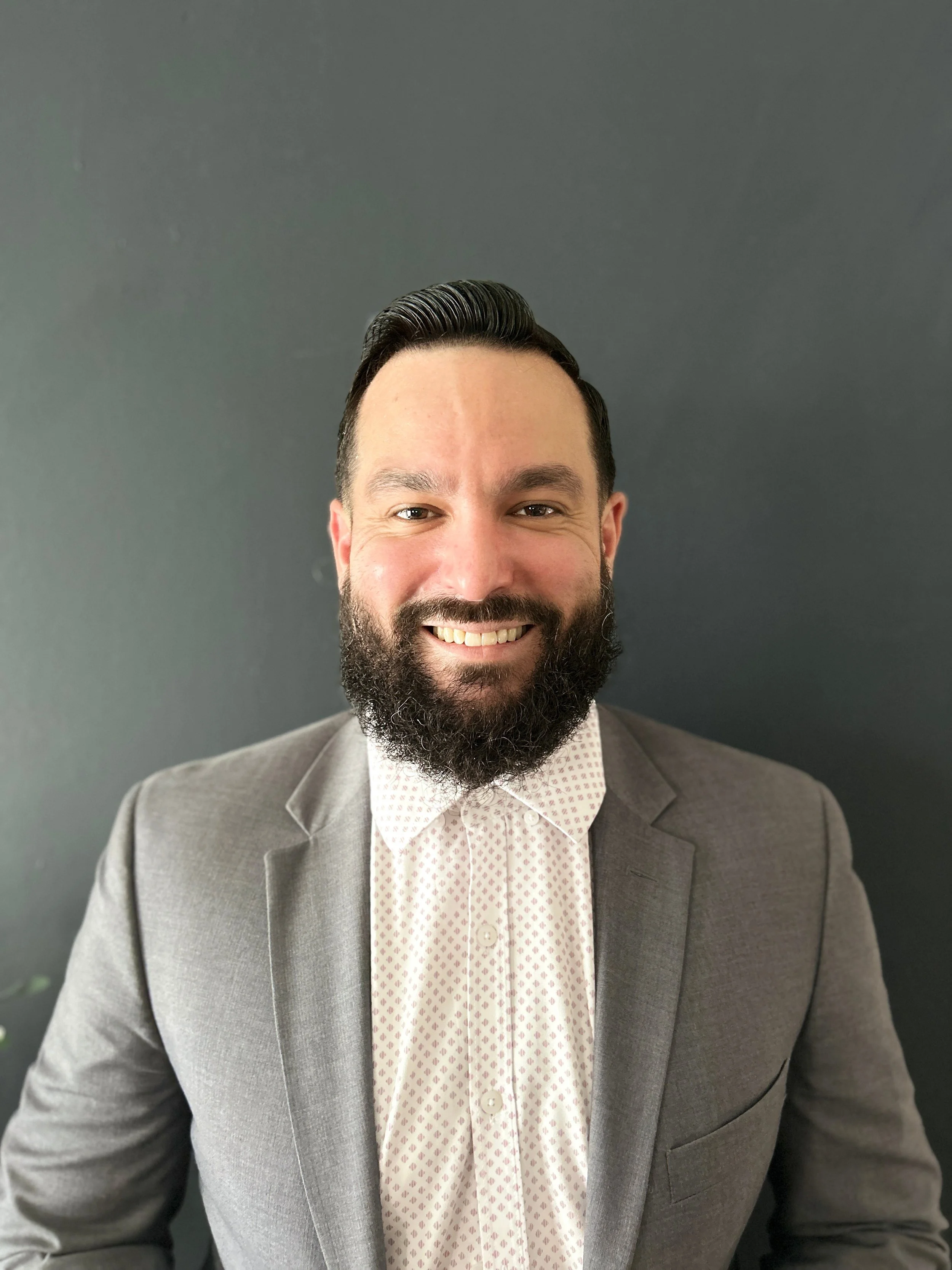A man with dark hair and a beard wearing a gray suit and a patterned white shirt smiling at the camera, against a dark gray background.