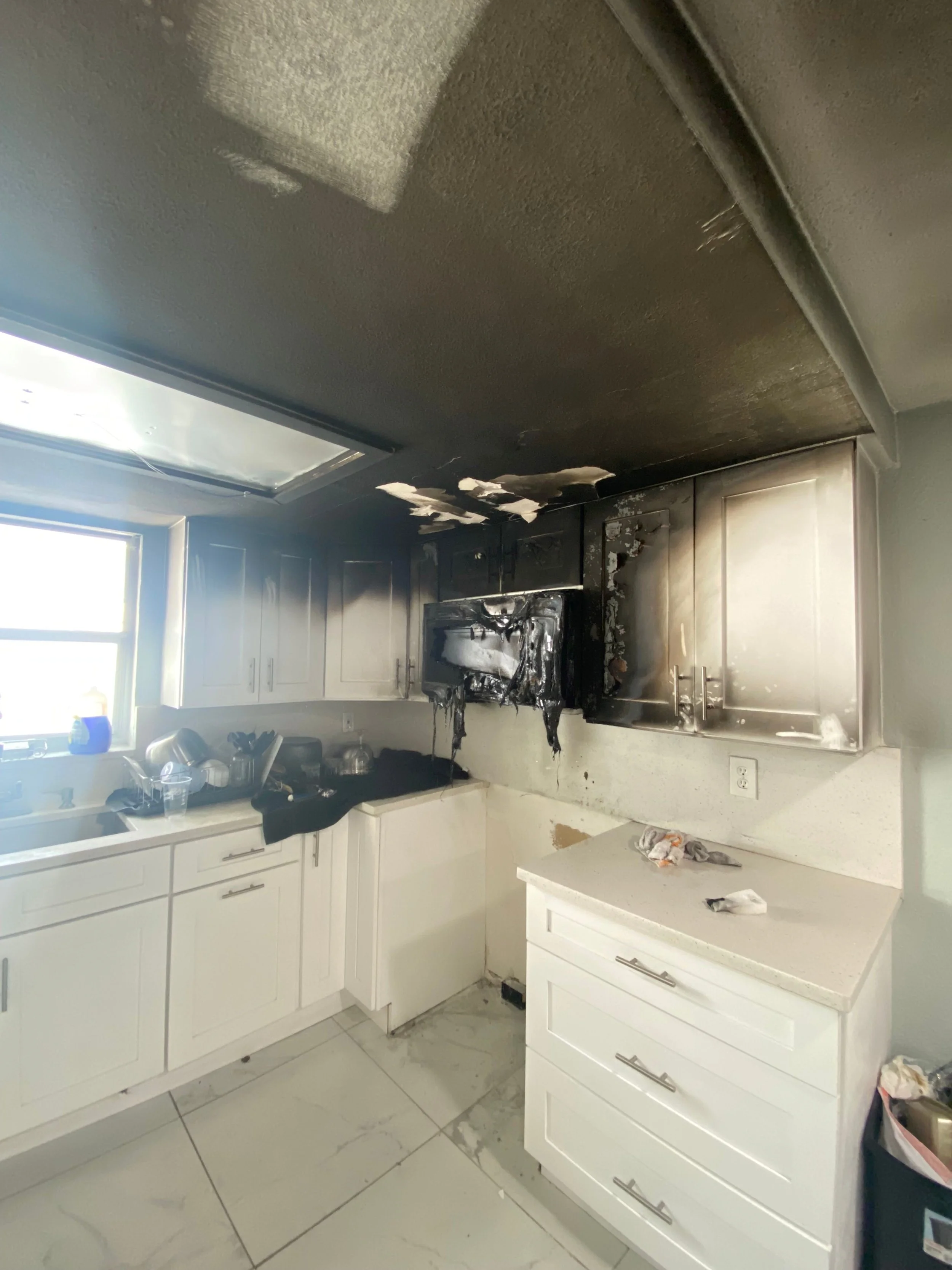 Kitchen with blackened and burnt cabinets and ceiling, indicating recent fire damage; some items on counters and floor.