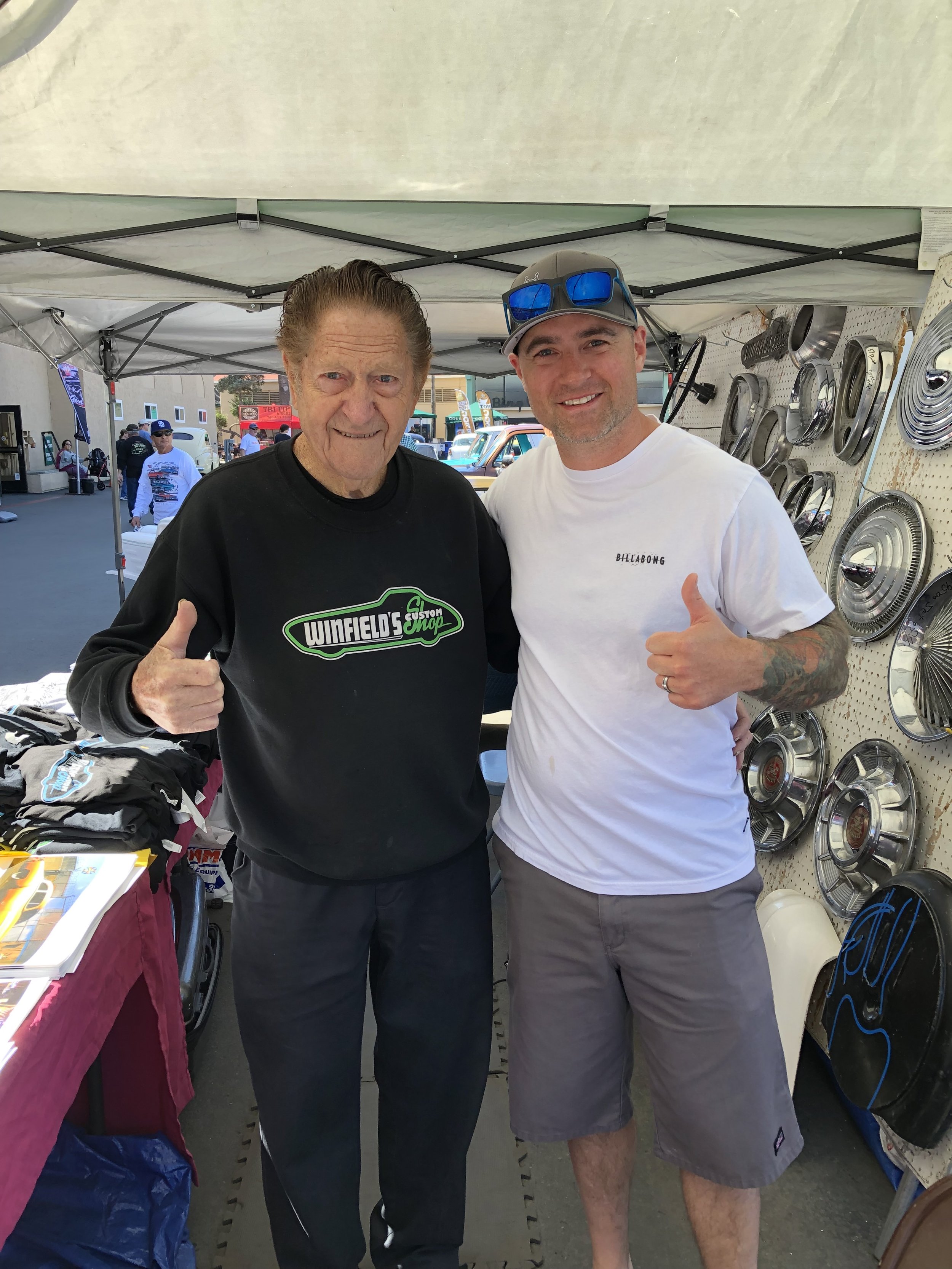 Two men standing together under a tent, giving thumbs up. One is wearing a black Winfield's Custom Shop sweatshirt, and the other is wearing a white T-shirt and sunglasses. They are at a car or auto part show, with hubcaps displayed behind them.