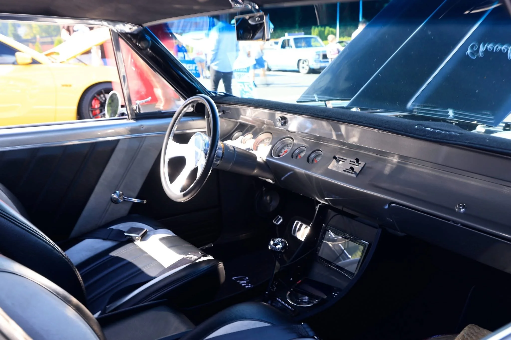 The interior of a vintage race car with a simple dashboard, a steering wheel, and racing seats, at an outdoor car event with other cars and people visible outside