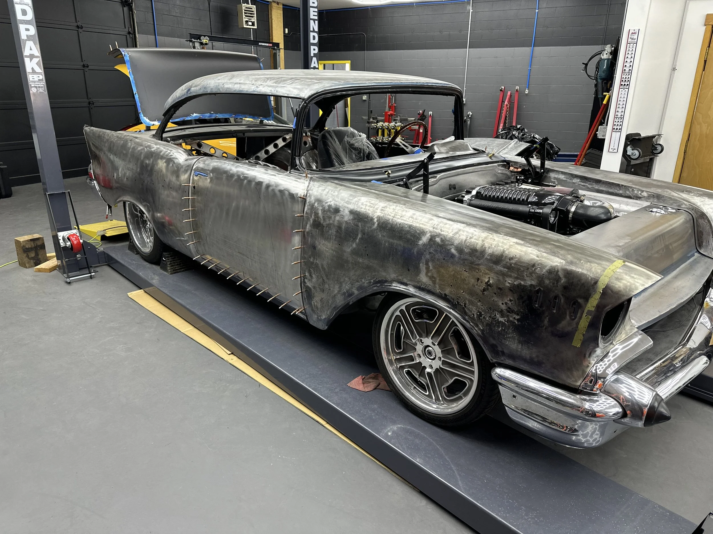A vintage car in a workshop undergoing restoration. The car's body is metal, partially sanded, with visible welding and unpainted surfaces. The front hood is open, exposing the engine. The car is mounted on a platform for work.