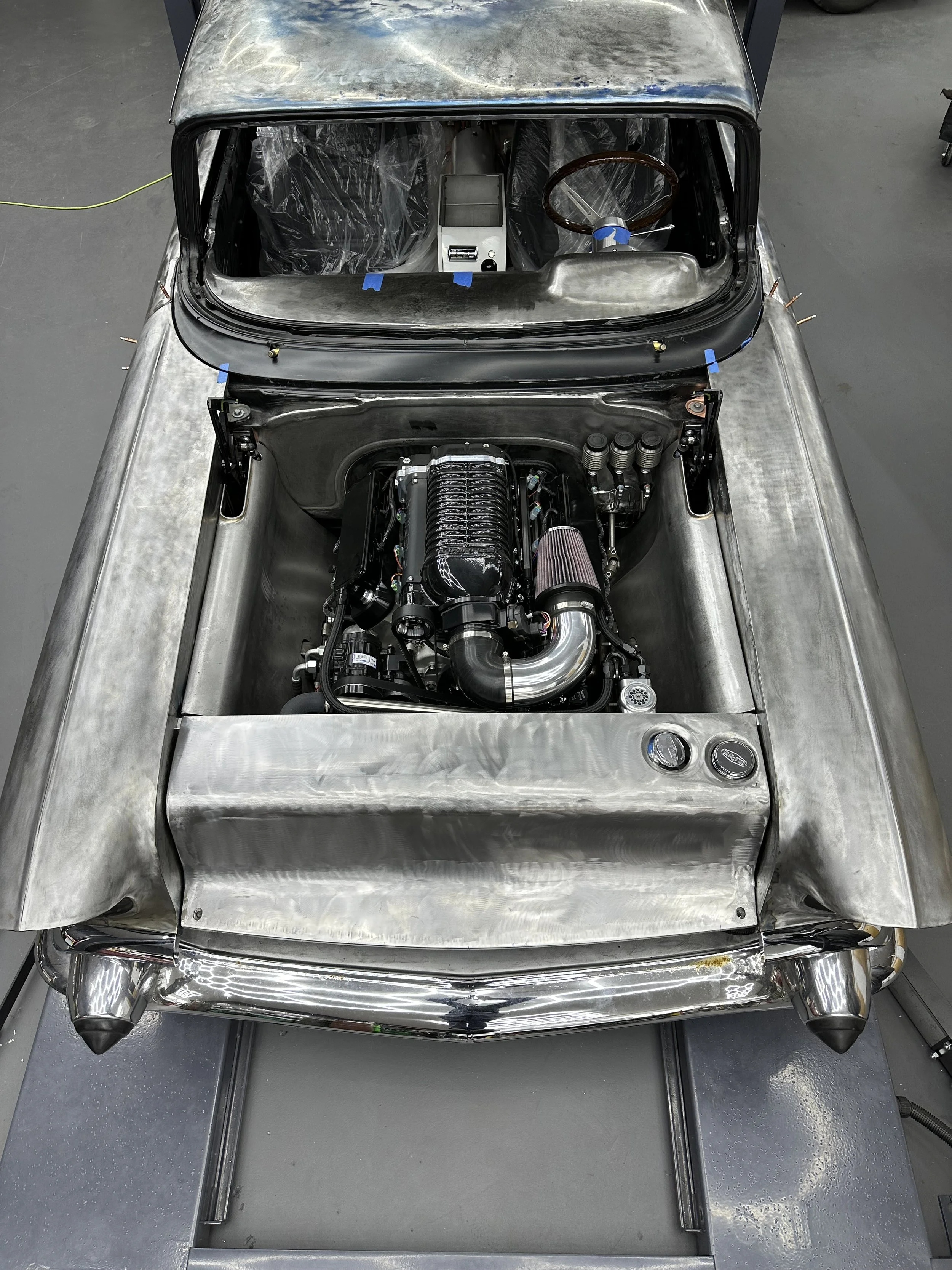 Uncarved vintage racing car with exposed metal body, showing engine and interior, in a workshop.