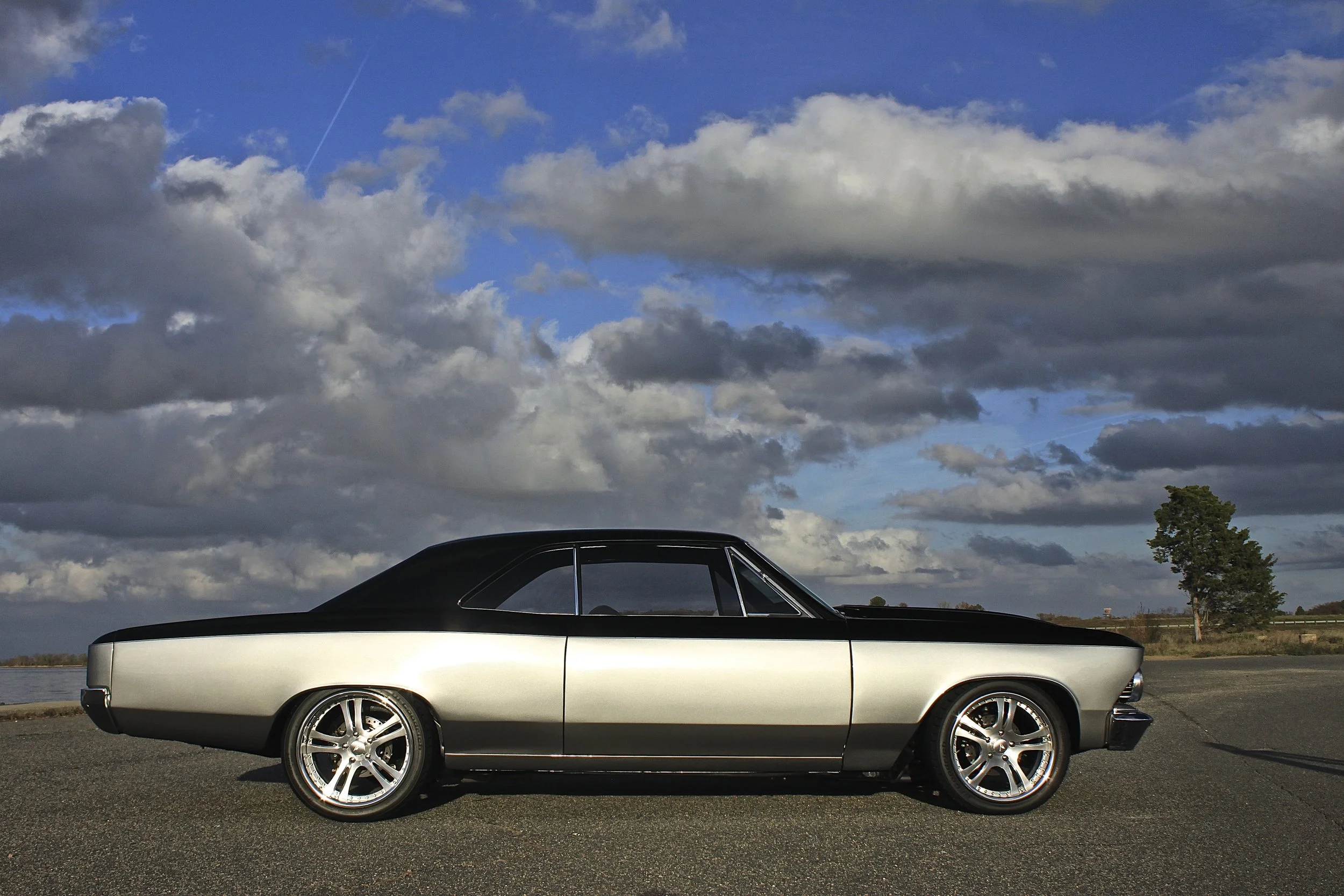 A vintage two-tone black and silver classic car parked on a paved surface outdoors under a cloudy sky.