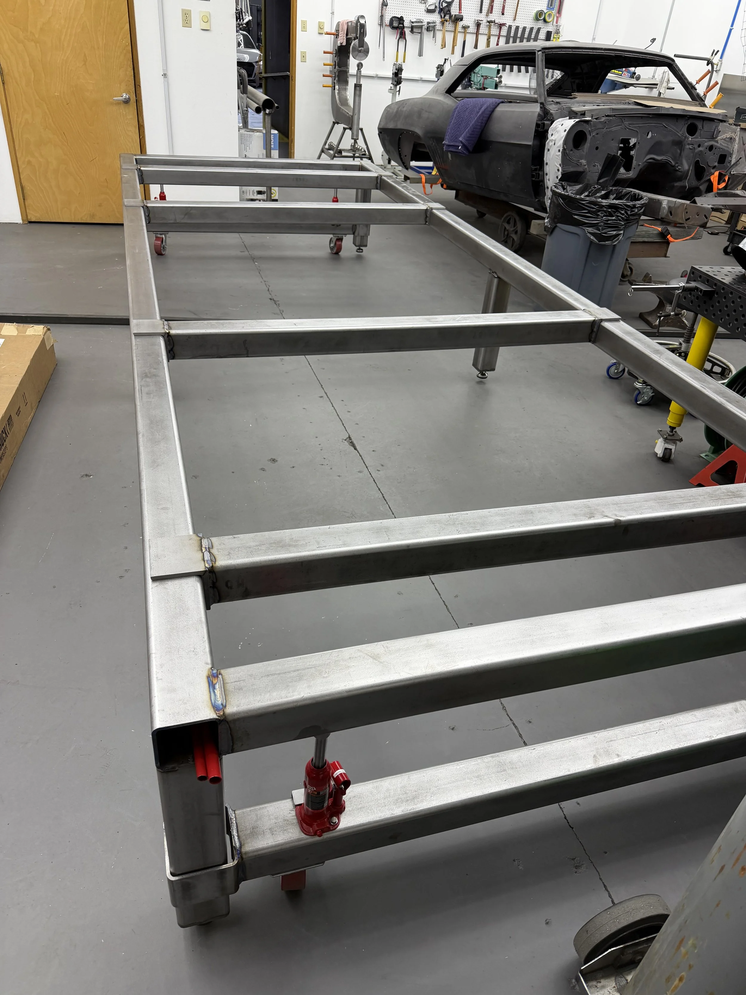 Unfinished metal vehicle frame in workshop with car body in background.