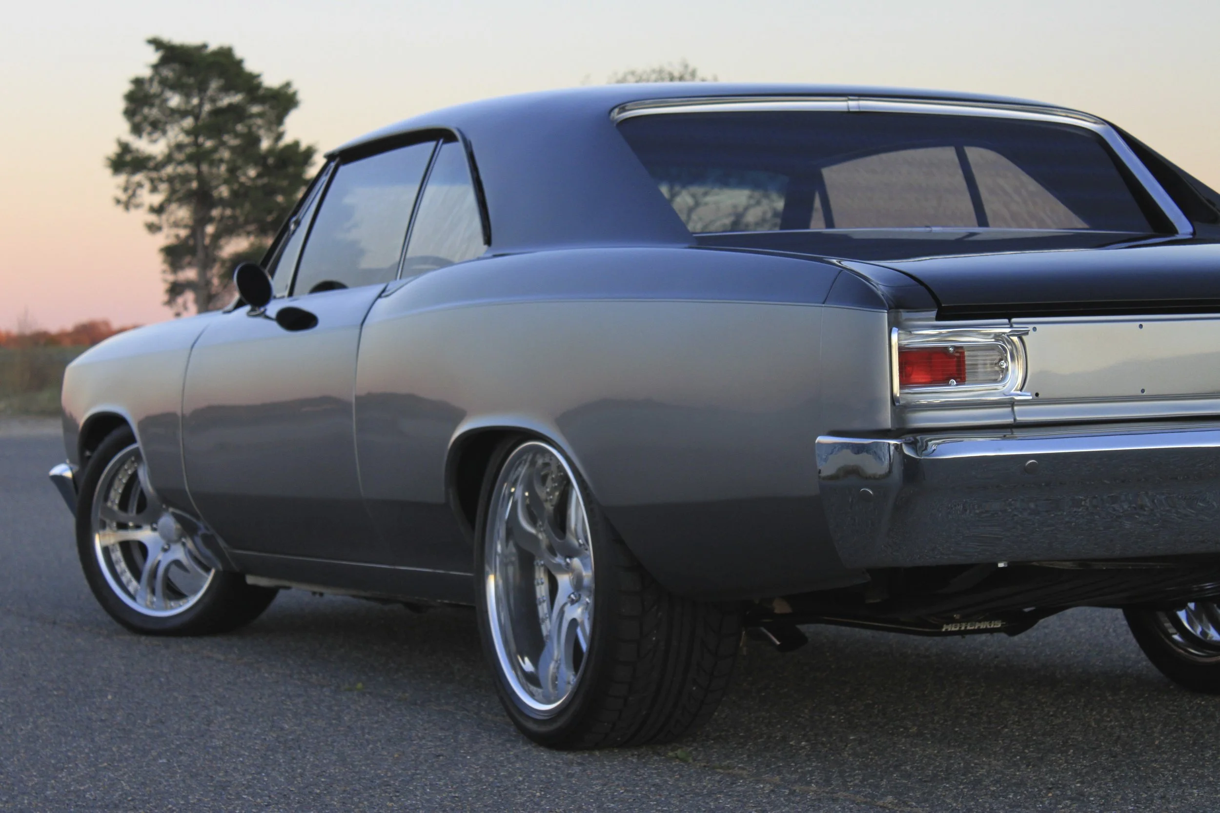 A classic two-tone gray and black vintage car with chrome accents, parked on a paved road during sunset with a tree and open sky in the background.