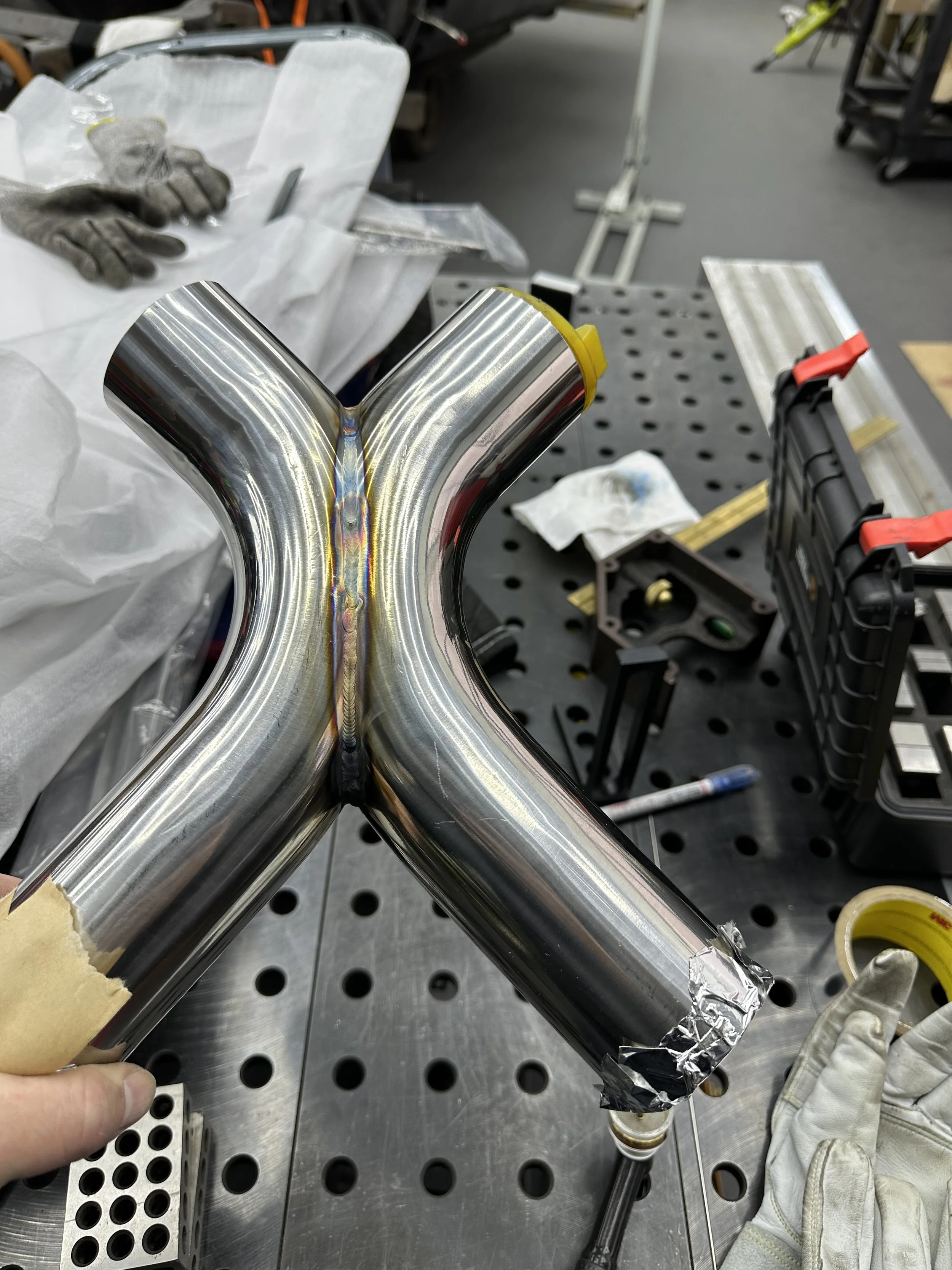 Close-up of a welded stainless steel Y-shaped pipe fitting on a workbench in a workshop.