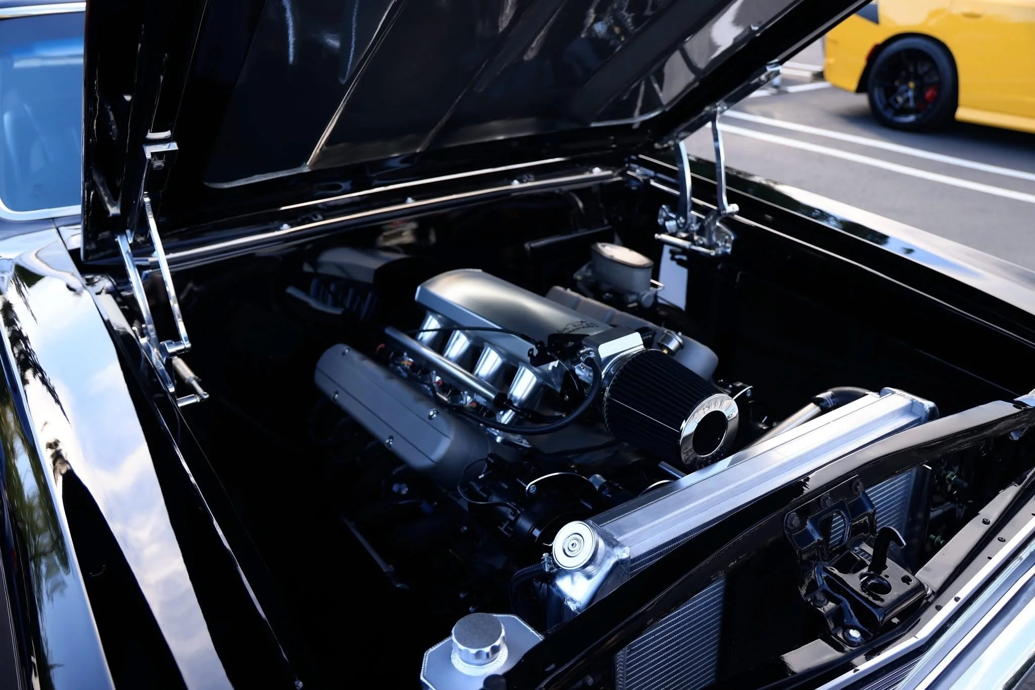 A close-up view of a car engine inside the engine bay of a black vehicle, with some yellow and blue cars visible in the background.