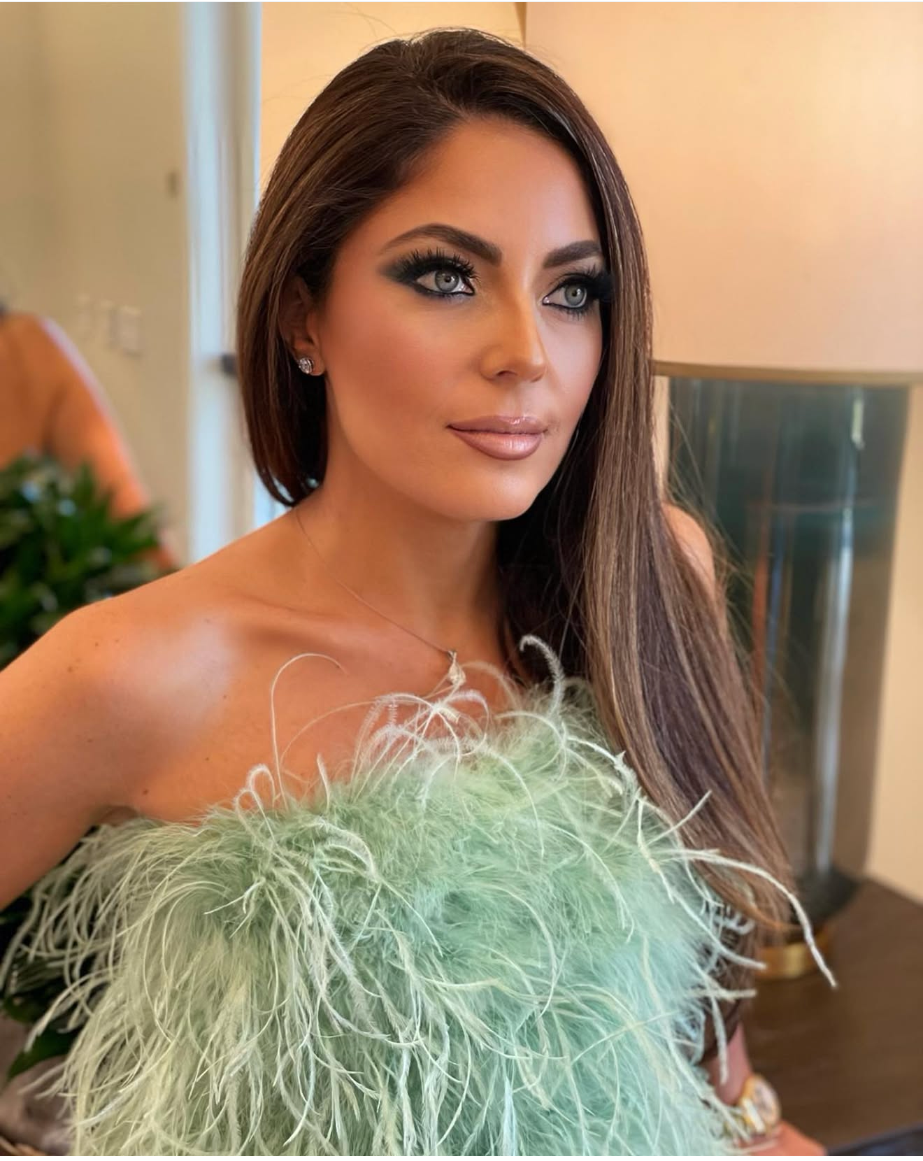 A woman with long brown hair and blue eyes, wearing heavy makeup and silver earrings, dressed in a strapless green feathered dress, sitting indoors with a blurred background.