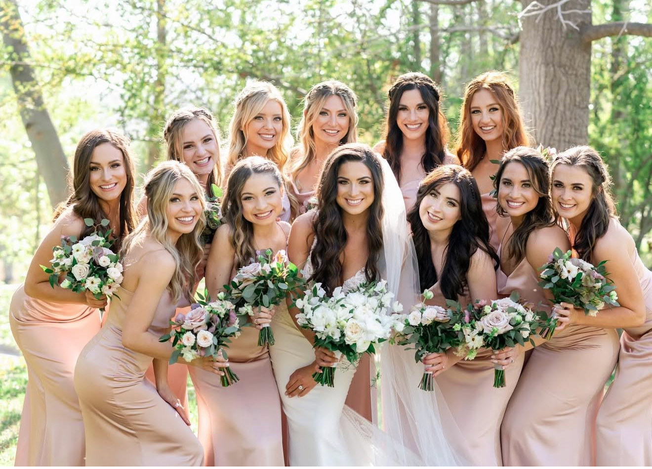 Group of women in light pink dresses holding bouquets of flowers outdoors for a wedding celebration.