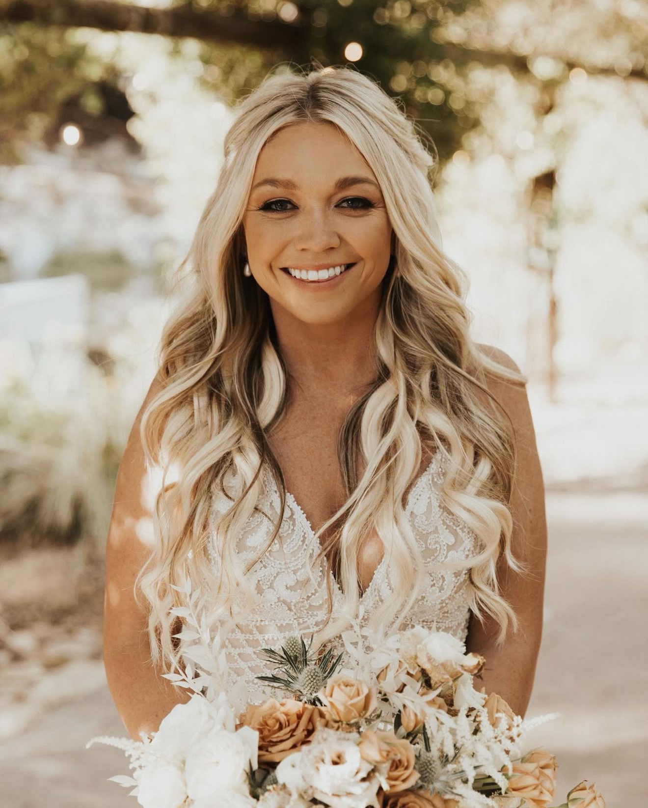 A woman with long, wavy blonde hair smiling in a wedding dress, holding a bouquet of peach, white, and cream flowers, standing outdoors with trees and blurred background.