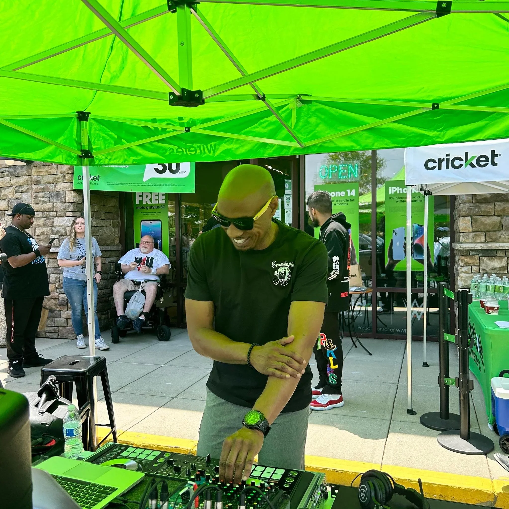 A man in a black shirt and gray pants smiling as he DJs at an event under a green tent outside a store with a stone facade, with people in the background.