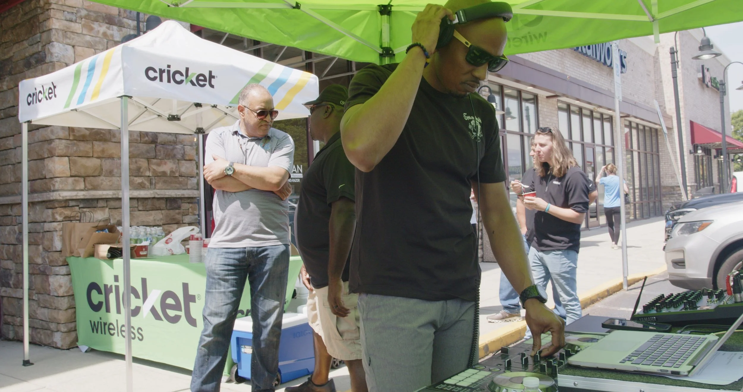 A man DJing outdoors under a green canopy at a Cricket Wireless promotional event. Several people are standing nearby, some using their phones, with a store and parked cars in the background.