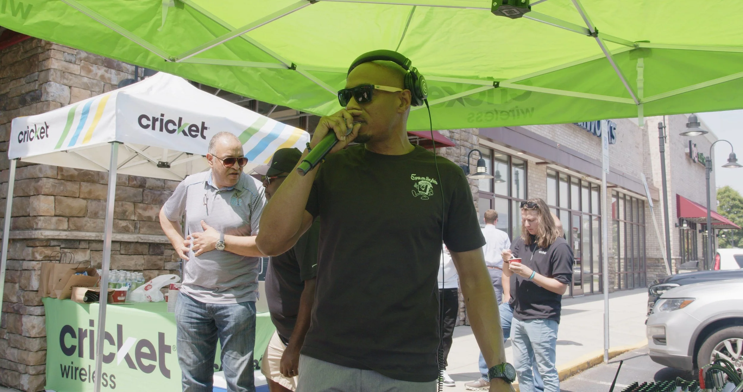 A man wearing sunglasses, headphones, and a black T-shirt is speaking into a microphone at an outdoor event under a green canopy. Several people are in the background near a booth with a white and green 