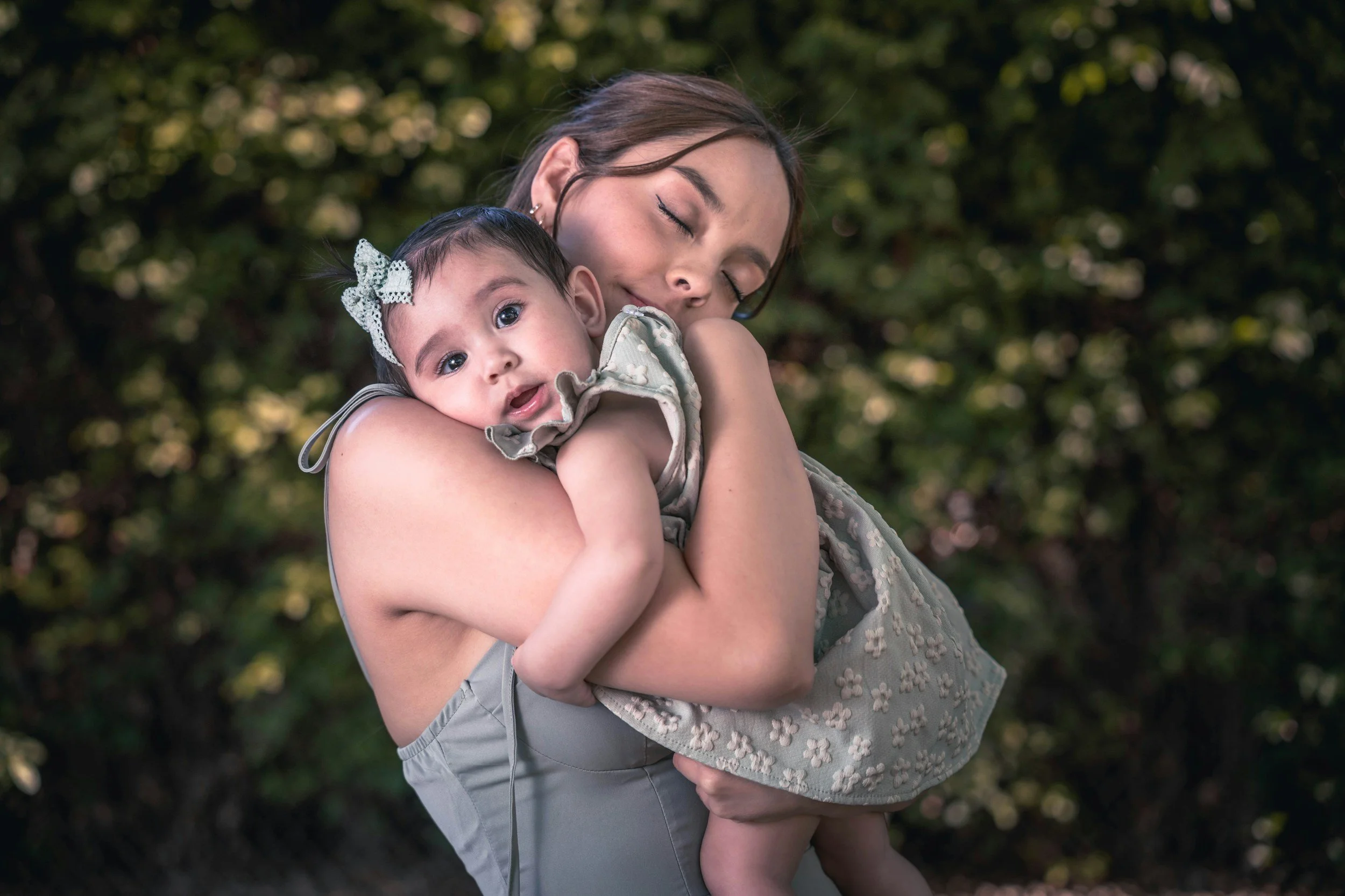 Una joven con los ojos cerrados abraza a una niña pequeña al aire libre, con un fondo de follaje verde.