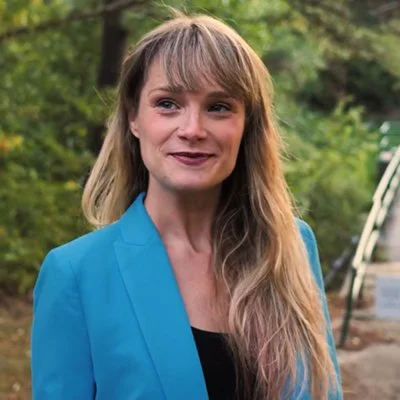 A woman with long blond hair and bangs wearing a blue blazer standing outdoors with trees in the background.