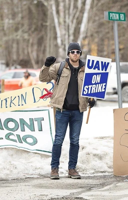 A man protesting with signs that say "UAW on Strike" and other messages, standing outdoors in cold weather with winter trees and parked cars in the background.