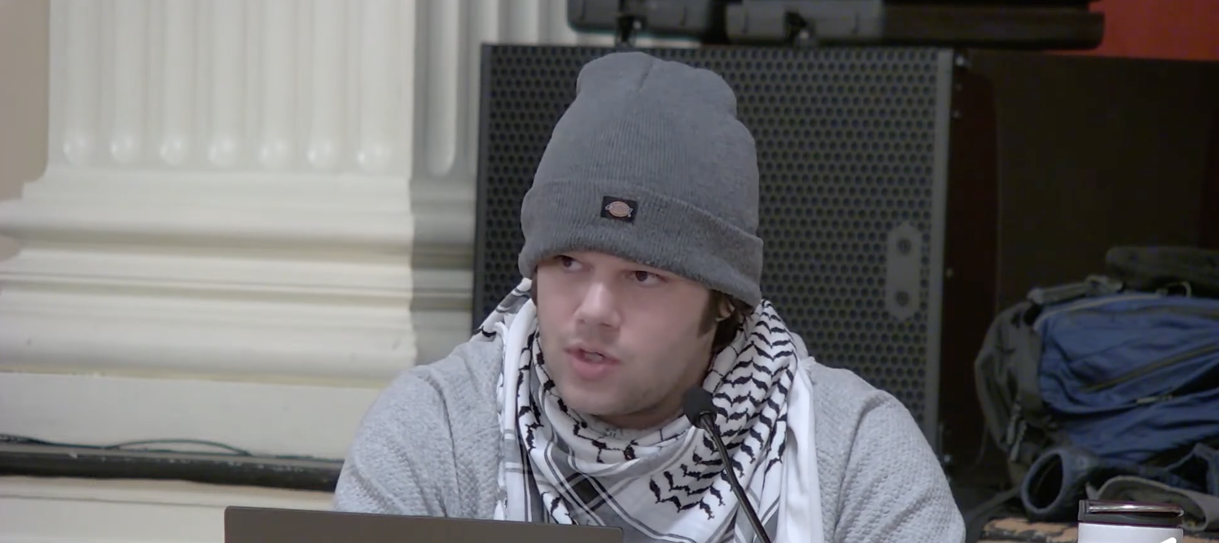 A young man wearing a gray beanie, gray sweater, and a black-and-white patterned scarf sits at a table with a microphone near his mouth, in a room with a white column, a black speaker, and a blue backpack.