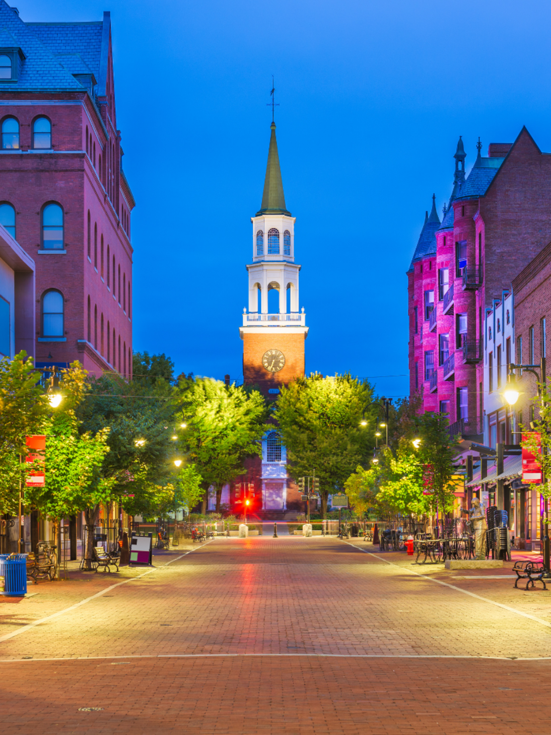 Empty brick-paved street in downtown with benches, lamp posts, trees, and shops, leading to a church tower with a clock at dusk.