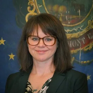 A woman with shoulder-length dark hair, glasses, and a black blazer smiling at the camera, standing in front of a decorative background with a blue section and gold stars.