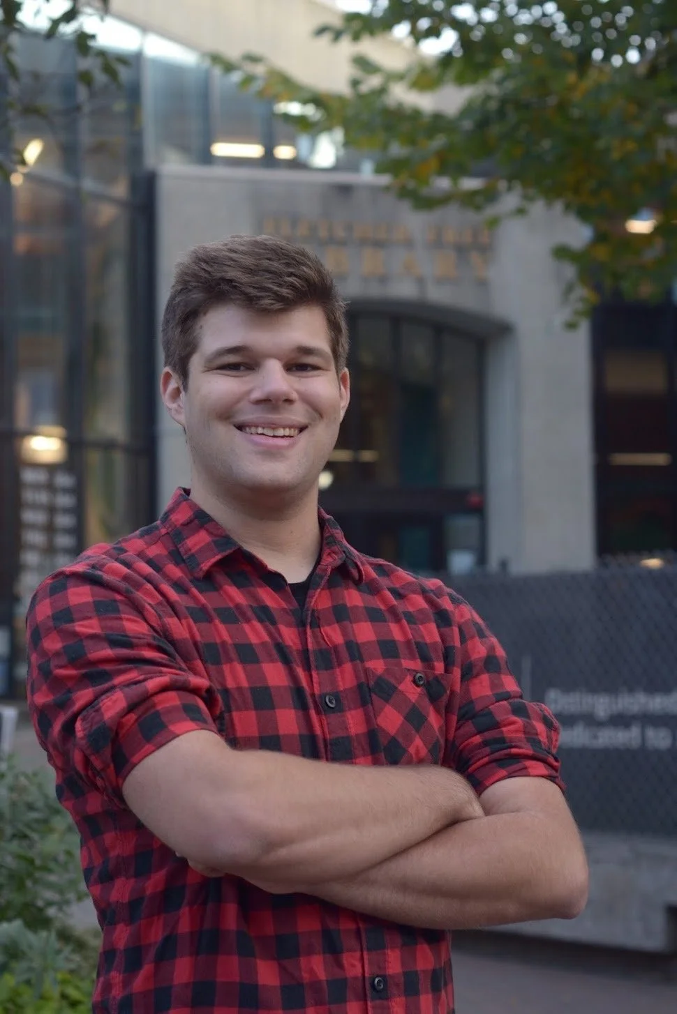Young man smiling with crossed arms, wearing a red and black plaid shirt, standing outdoors in front of a building and trees.
