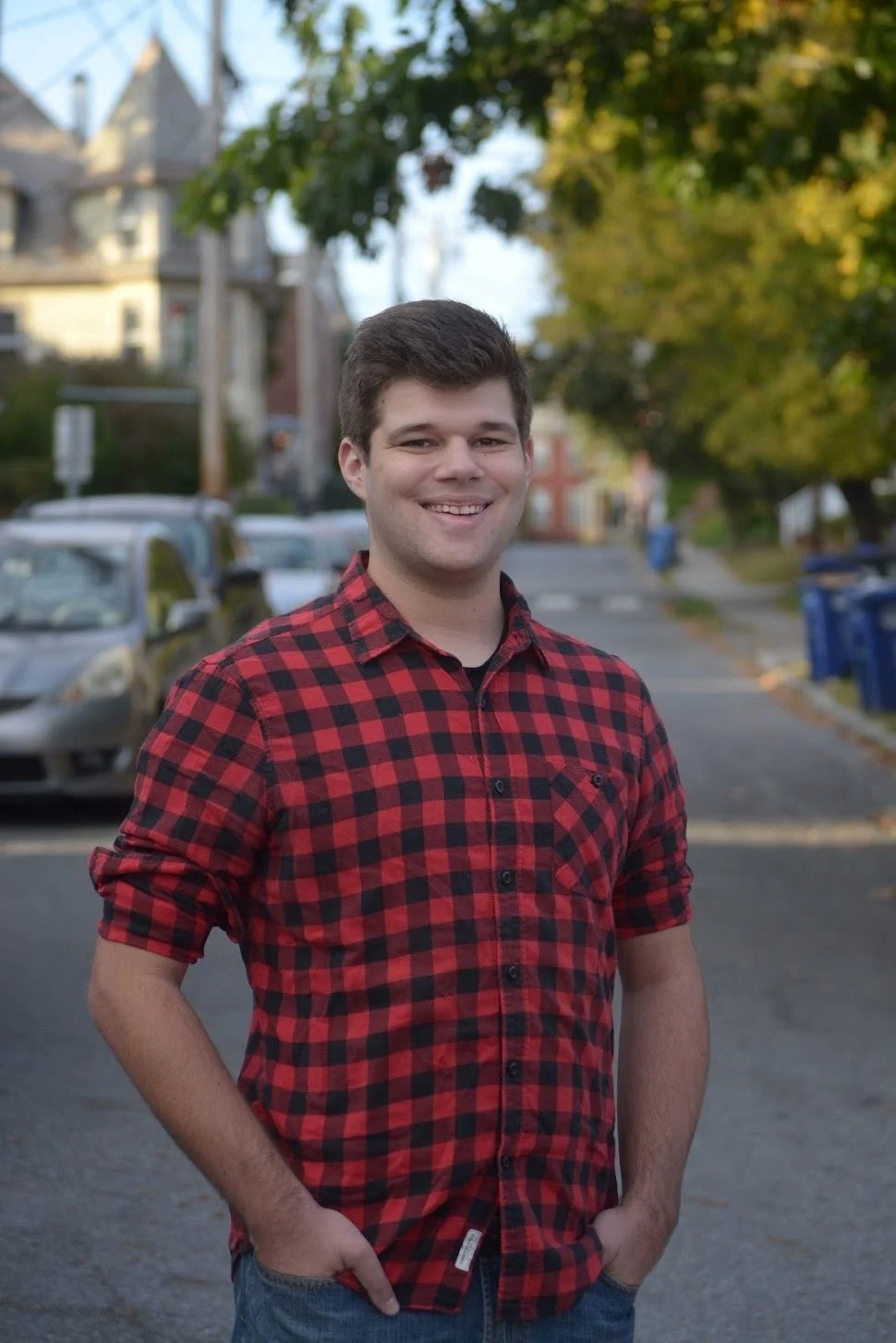 A young man smiling outdoors on a residential street during daylight, wearing a red and black plaid shirt with hands in pockets.