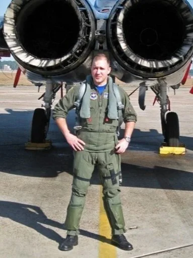 A man in military flight suit standing in front of a jet aircraft with large jet engines at an airfield.