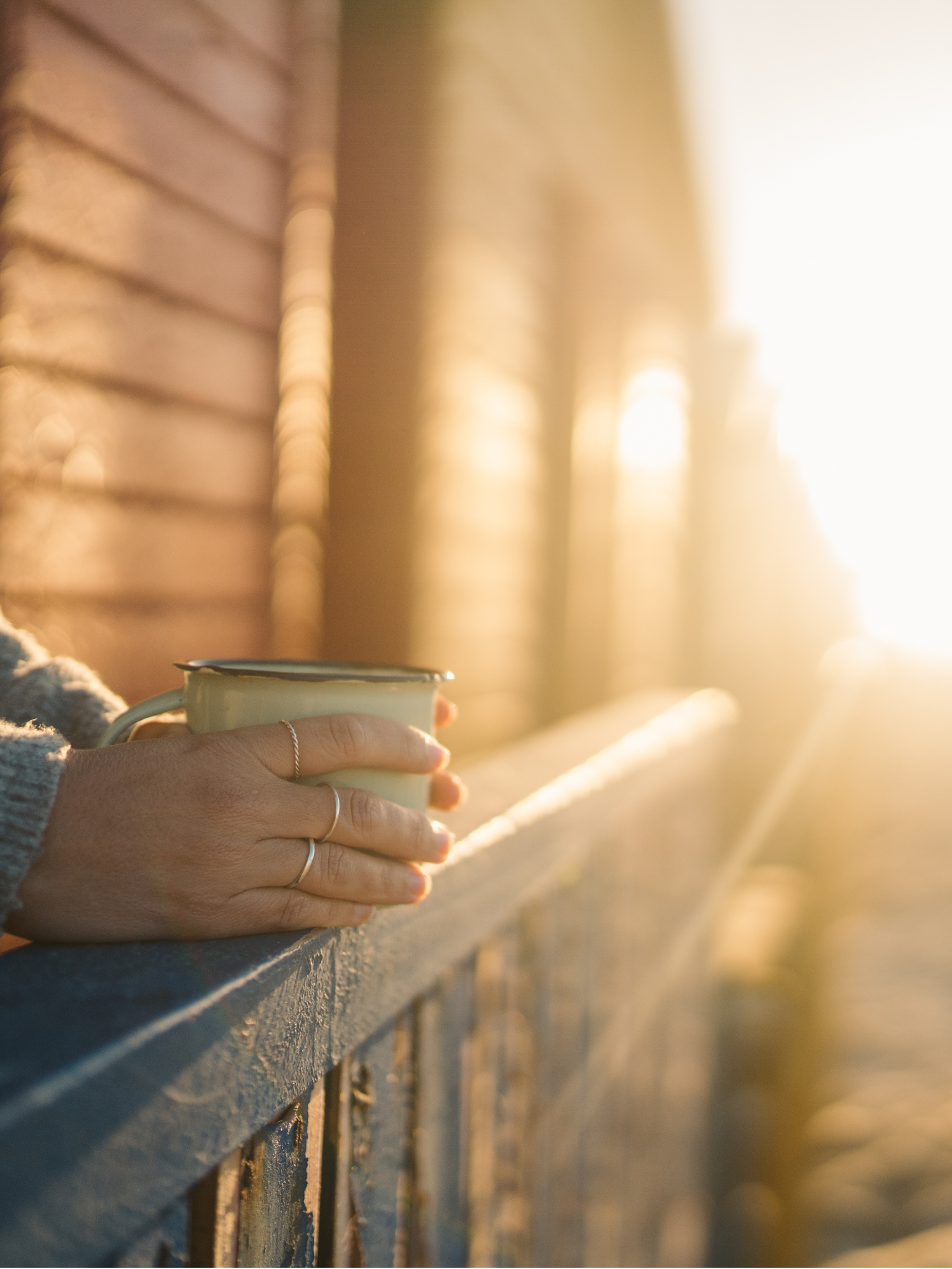 A person with rings on their fingers holding a mug, resting their hands on a wooden railing during sunset or sunrise. Calm counselling sensation