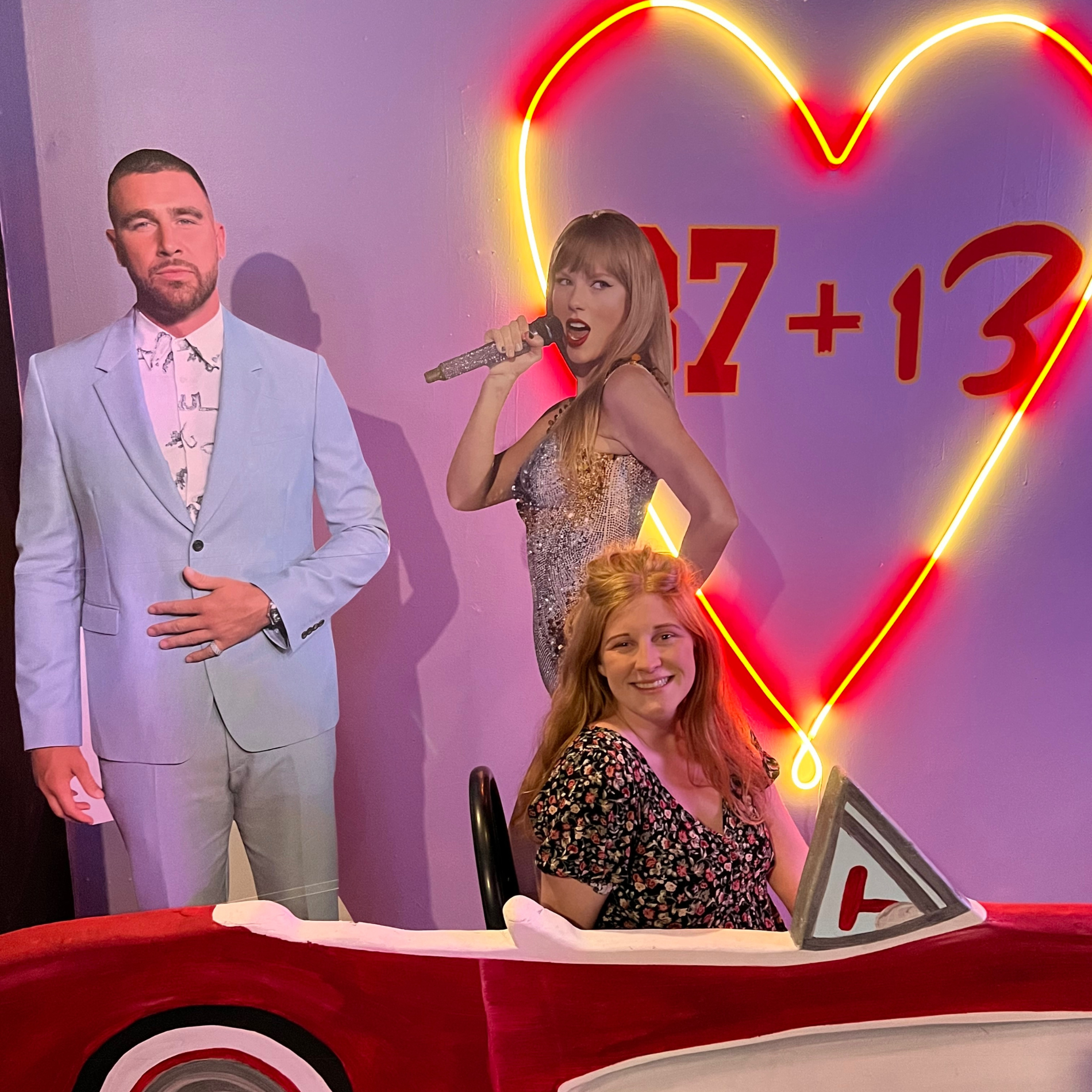 A photo of three people, two women and one man, posing in front of a wall with a neon heart and a math equation '27 + 13'. The woman in the foreground is sitting in a red paper car as if driving, smiling at the camera. The woman in the background is holding a microphone, wearing a sparkly dress, and appears to be singing or speaking. The man is dressed in a light-colored suit and stands with his hand over his stomach, looking at the camera.